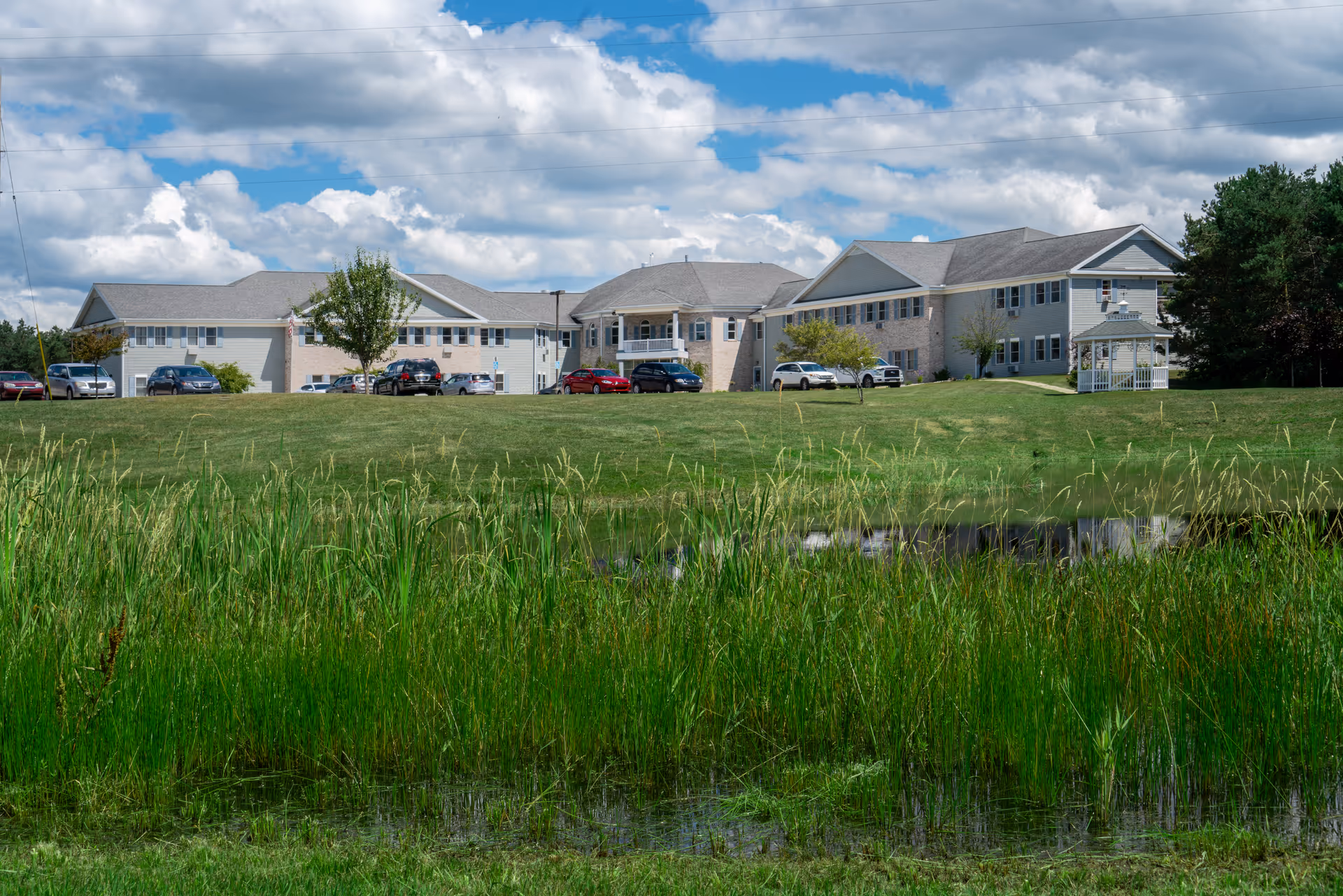 Two-story senior living building with parked cars and a gazebo behind a grassy lawn and pond under a partly cloudy sky.