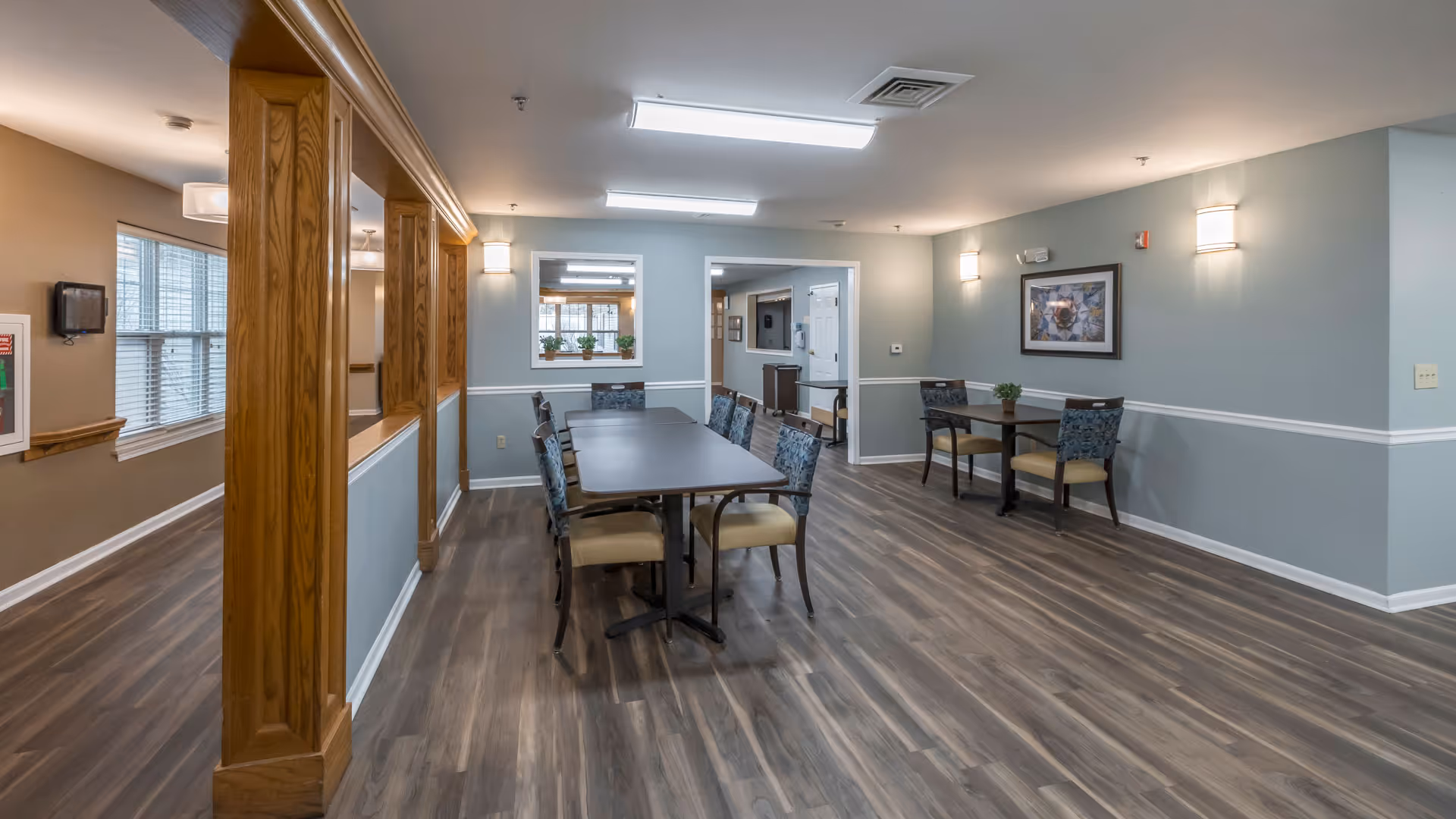 A spacious dining area in a senior living facility with wooden flooring, light blue and beige walls, and multiple tables with chairs. The room is well-lit with ceiling lights and wall sconces. There are windows and framed artwork on the walls, and a wooden partition separates this area from an adjacent hallway.