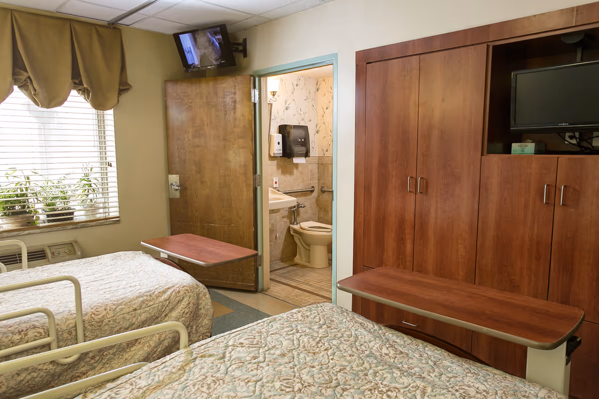 Two-bed senior care bedroom with overbed tables, wooden cabinetry, a wall-mounted TV, and an open door to a small bathroom.