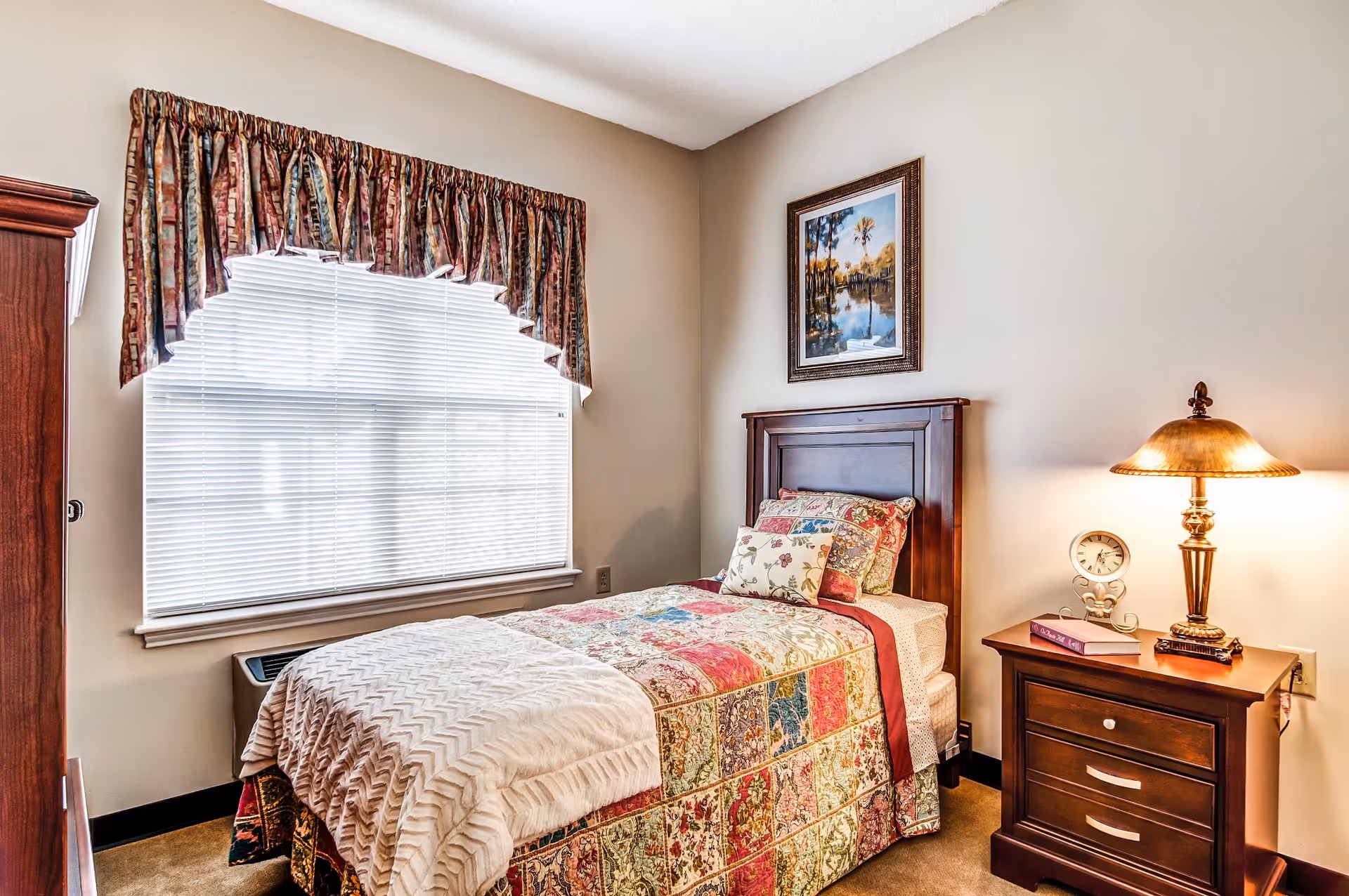 Cozy bedroom with a single bed dressed in a patchwork quilt, wooden headboard, nightstand with a lamp, and a window with blinds.