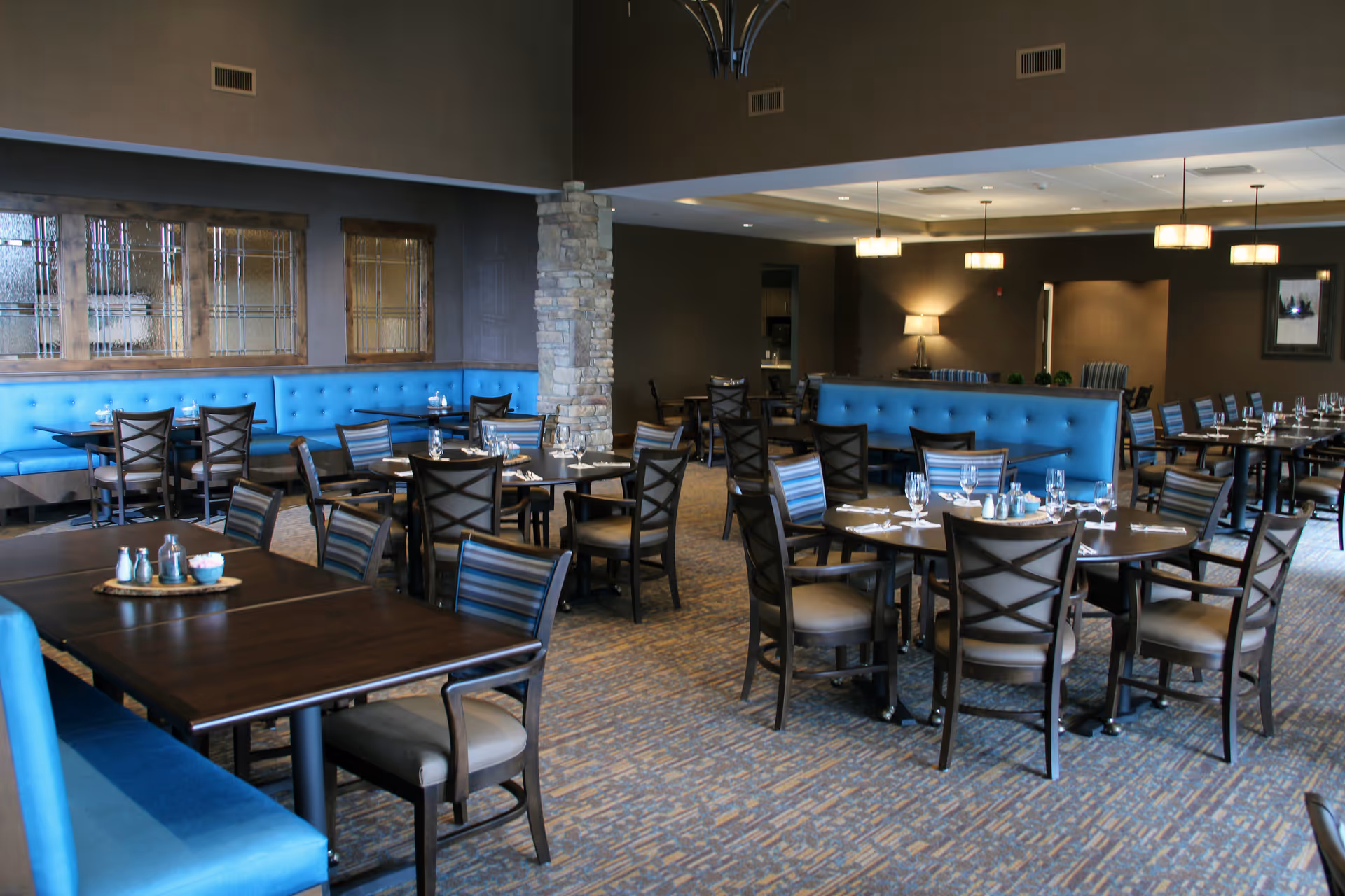 A spacious dining room in a senior living community featuring multiple dark wood tables and chairs with beige cushions. The room has blue upholstered bench seating along the walls, stone pillars, and warm lighting from ceiling fixtures and a table lamp. Tables are set with glasses, napkins, and condiments.