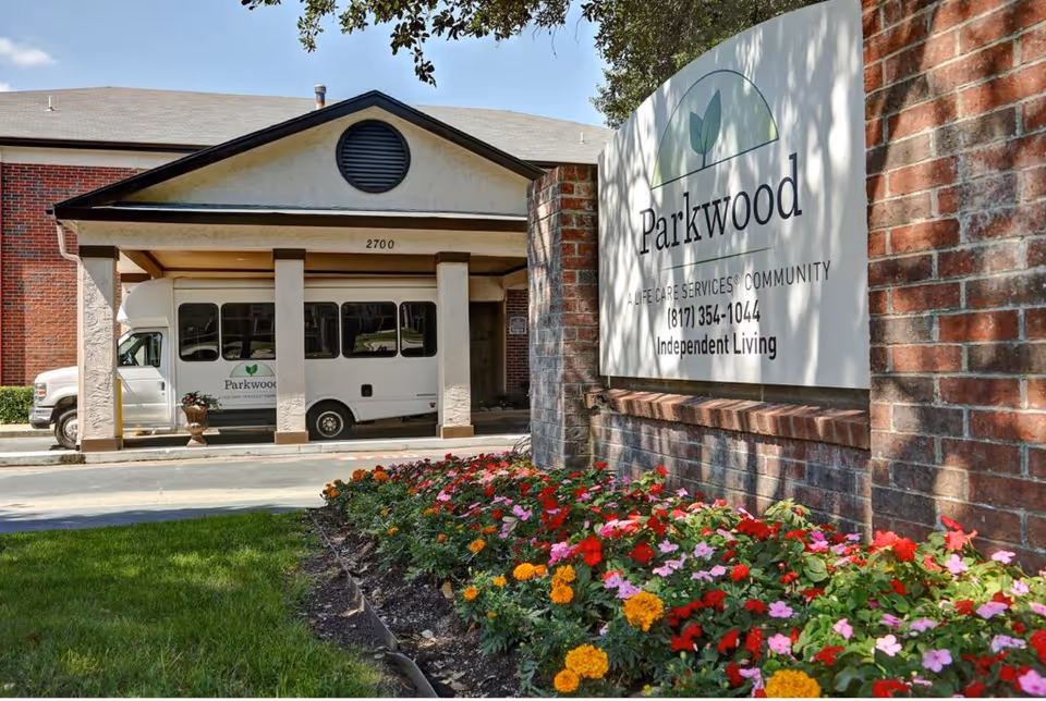 Entrance of Parkwood Healthcare facility showing a covered driveway with a white shuttle bus parked under it. A large brick sign with the Parkwood logo and contact information is visible next to a flower bed with colorful flowers in front of the building.