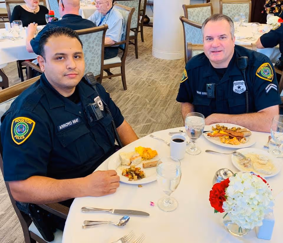 Two police officers in Houston Police uniforms sitting at a round dining table with plates of breakfast food, including eggs, bacon, and biscuits. The setting appears to be a dining room with other people seated at tables in the background. A small floral centerpiece with white and red flowers is on the table.