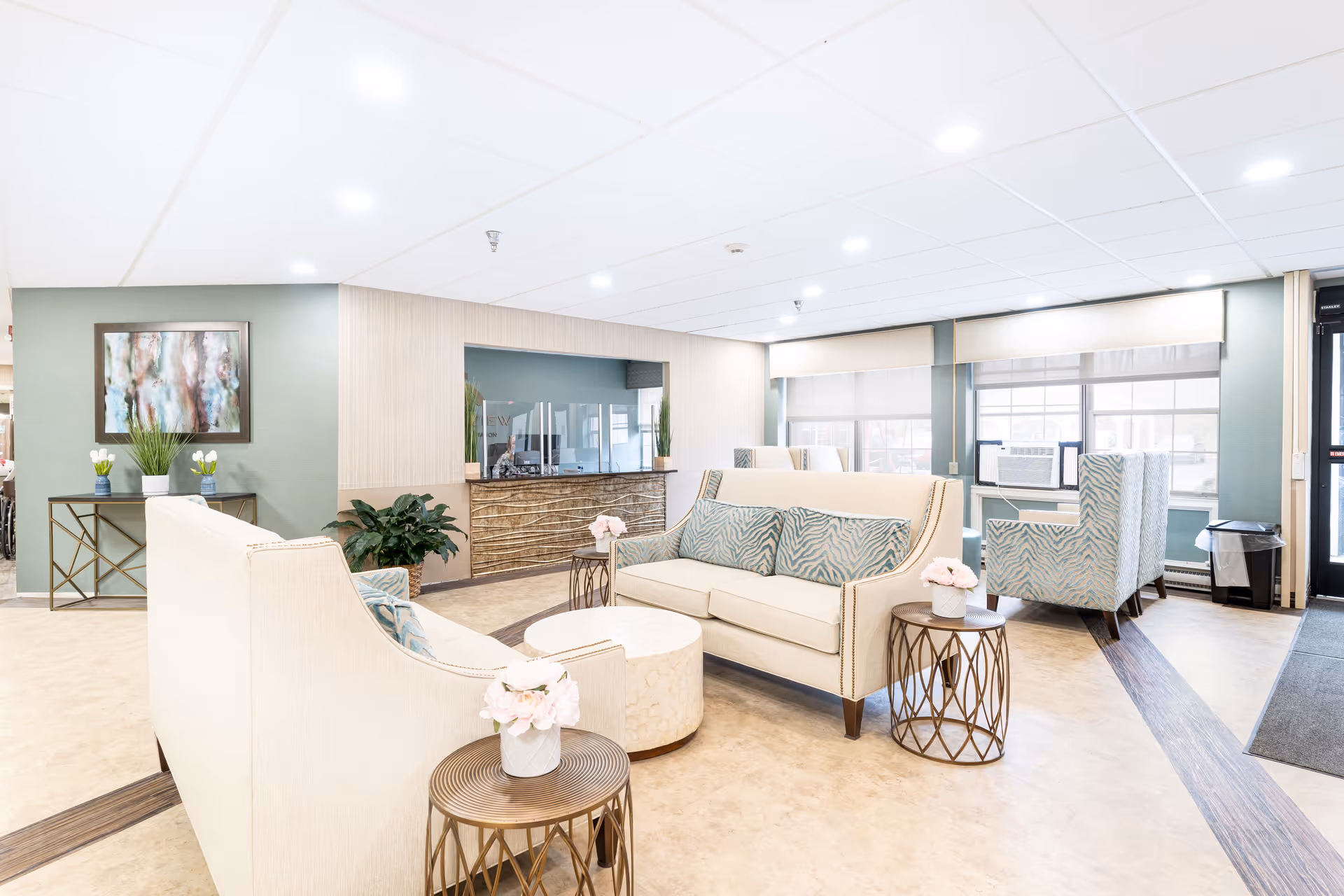 A bright and spacious nursing facility common area with cream-colored sofas and armchairs featuring blue patterned cushions. There are small round tables with flower vases, large windows with blinds, a reception desk with a glass partition, and decorative plants and artwork on the walls.