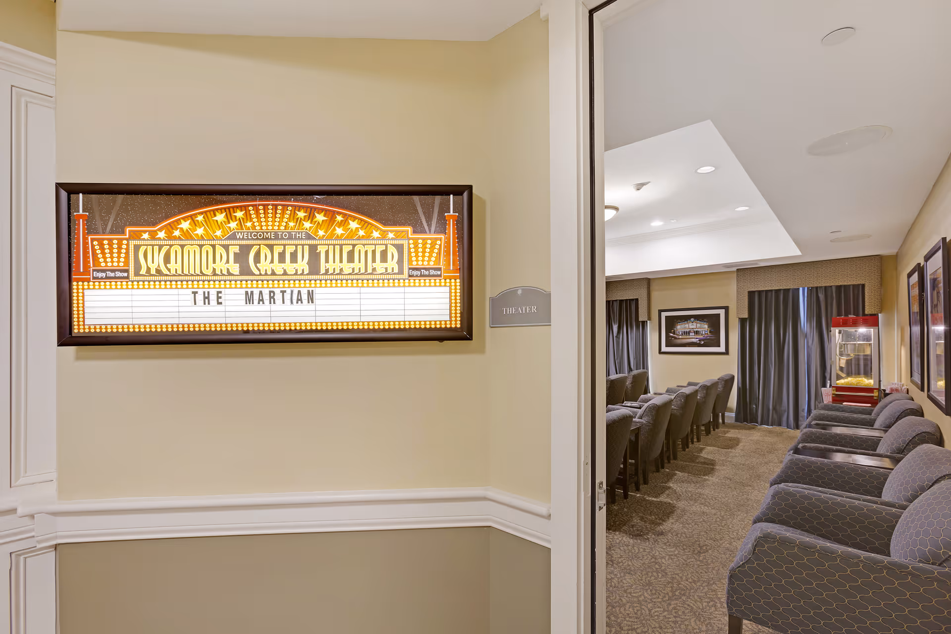 Entrance to a small theater room with a lighted marquee sign reading 'Welcome to the Sycamore Creek Theater' and 'The Martian'. Inside, rows of cushioned chairs face a screen, with a popcorn machine visible in the back corner.