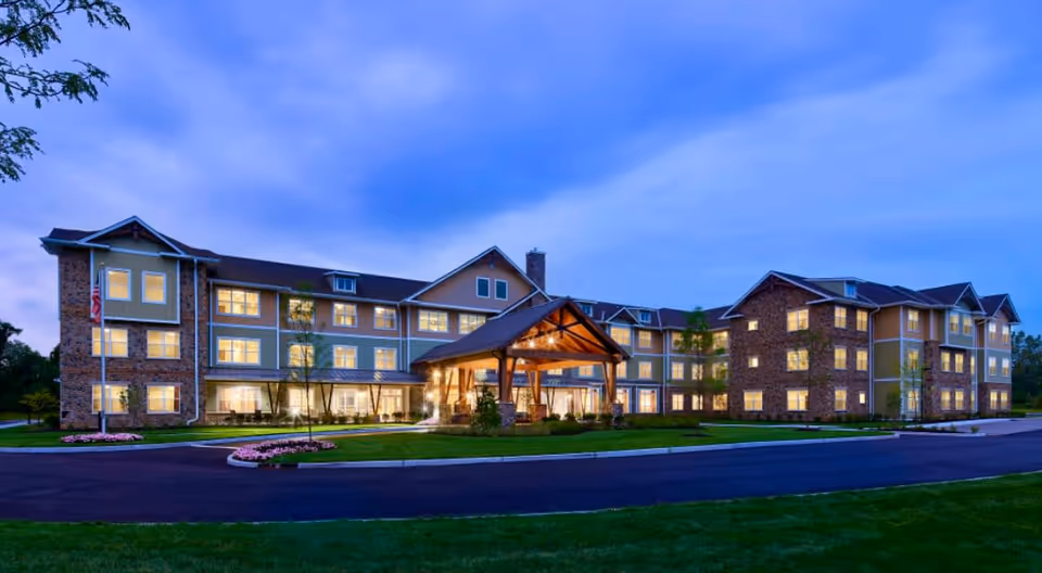 Exterior view of The Chelsea at Greenburgh senior living facility at dusk, showing a large multi-story building with many lit windows, a covered entrance with wooden beams, landscaped grounds, and a driveway in front.