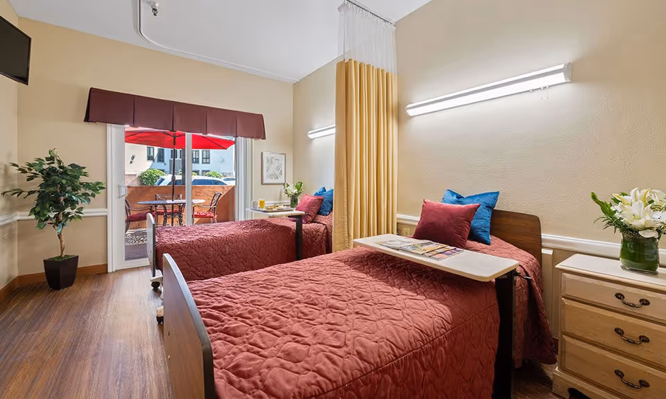 A senior living bedroom with two beds covered in red quilts, a nightstand, plants, and a sliding door opening to a patio with a red umbrella.
