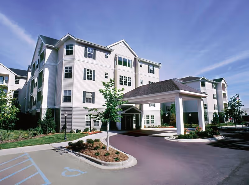 Exterior view of a multi-story senior living facility building with white siding, multiple windows, and a covered entrance driveway. There are small trees and landscaped areas around the building, and a clear blue sky overhead.