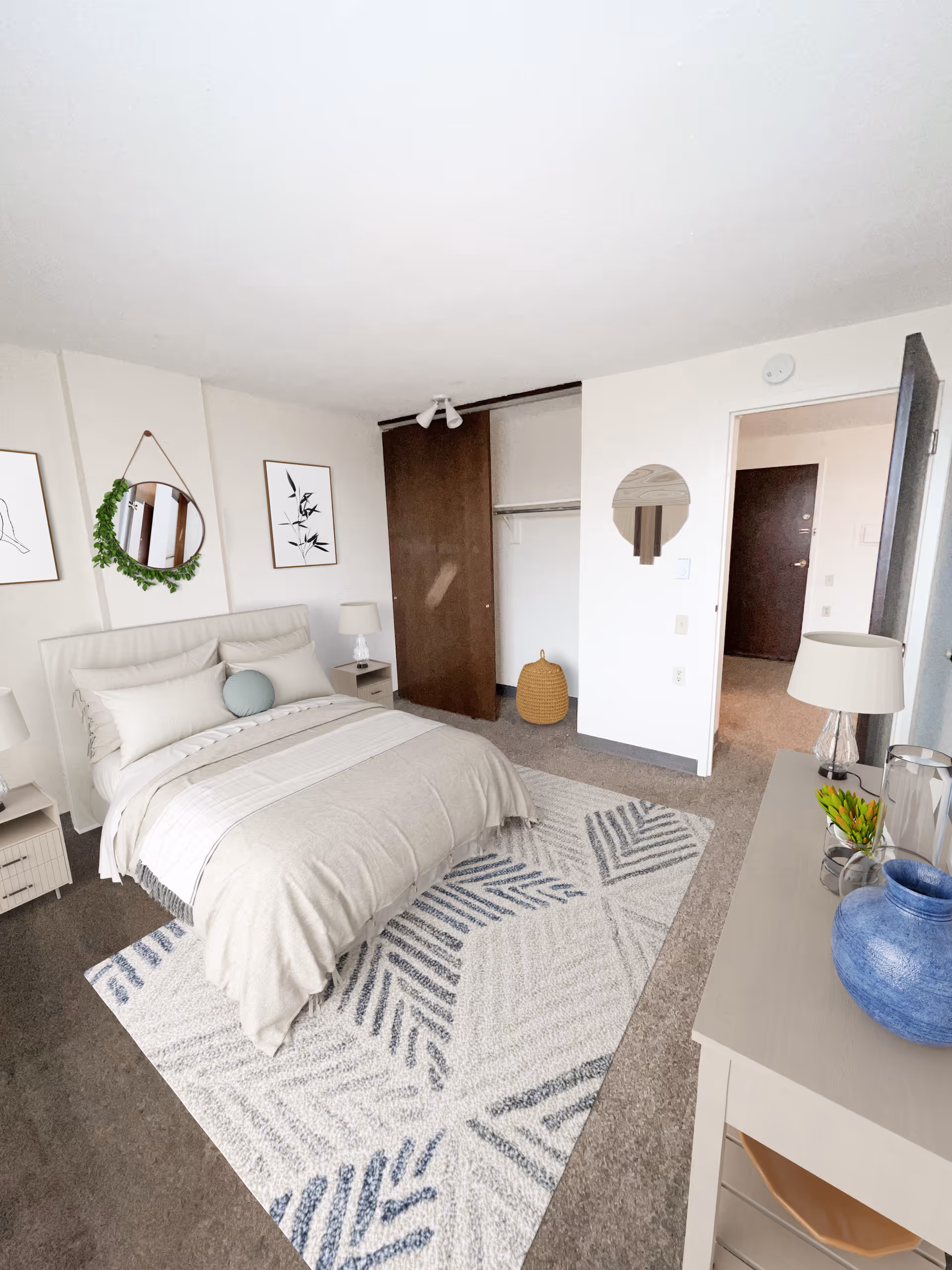 A neatly arranged bedroom with a bed covered in beige and white bedding, two bedside tables with lamps, a patterned rug on the floor, a dresser with decorative items including a blue vase and a small plant, a round mirror on the wall, and an open closet with a wooden sliding door.