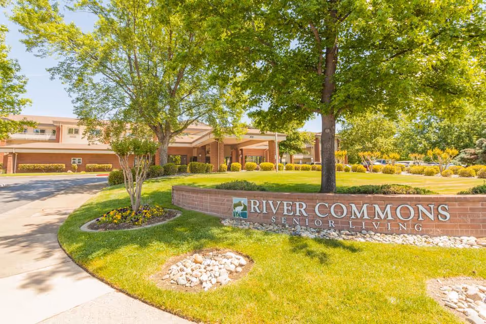 Exterior view of River Commons Senior Living facility with a brick sign in front, green grass, trees, and a driveway leading to the building entrance.