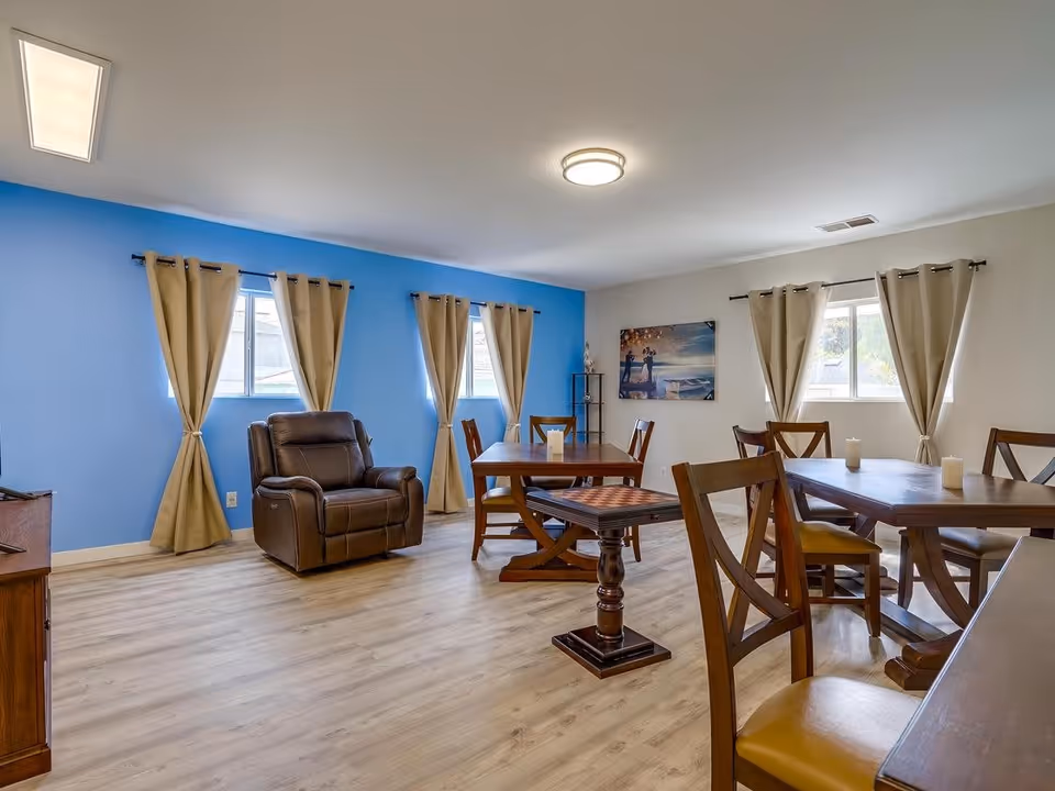 A bright room with light wood flooring and beige curtains on windows. The walls are painted blue and white. The room contains several wooden tables and chairs, a brown leather recliner chair, and a small wooden table with a checkerboard pattern on top. There is a painting on the wall and a small shelf in the corner.