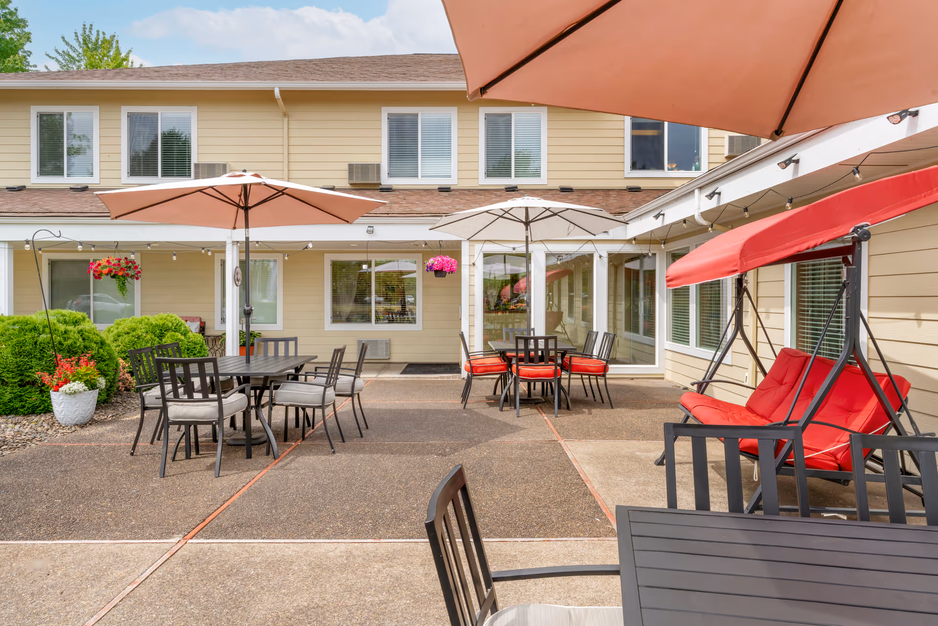 Outdoor patio area at Brookdale Heritage Plaza featuring multiple tables with umbrellas, chairs with cushions, a red cushioned swing, hanging flower baskets, and a beige two-story building in the background.