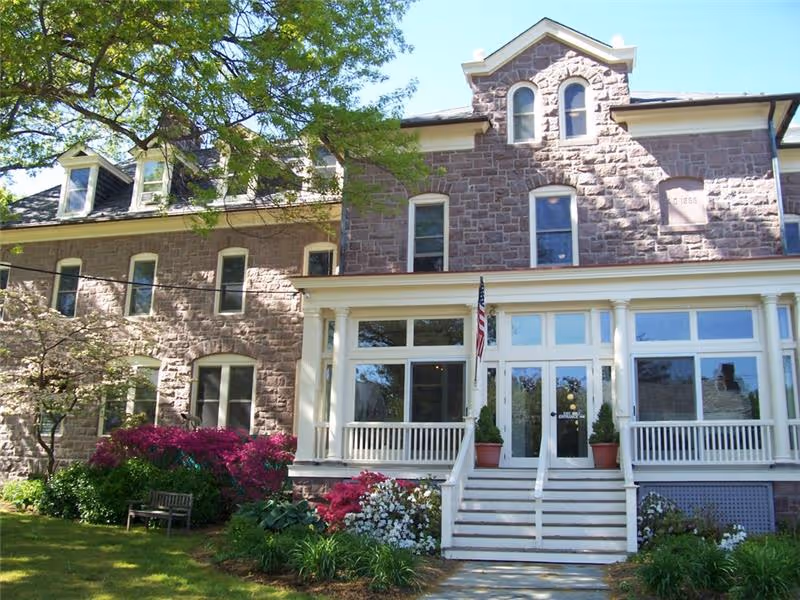 Front exterior view of a large stone building with multiple windows and a white porch with stairs leading up to the entrance. There are flowering bushes and green plants in the garden in front of the building, and a tree partially shading the area. An American flag is displayed near the entrance.