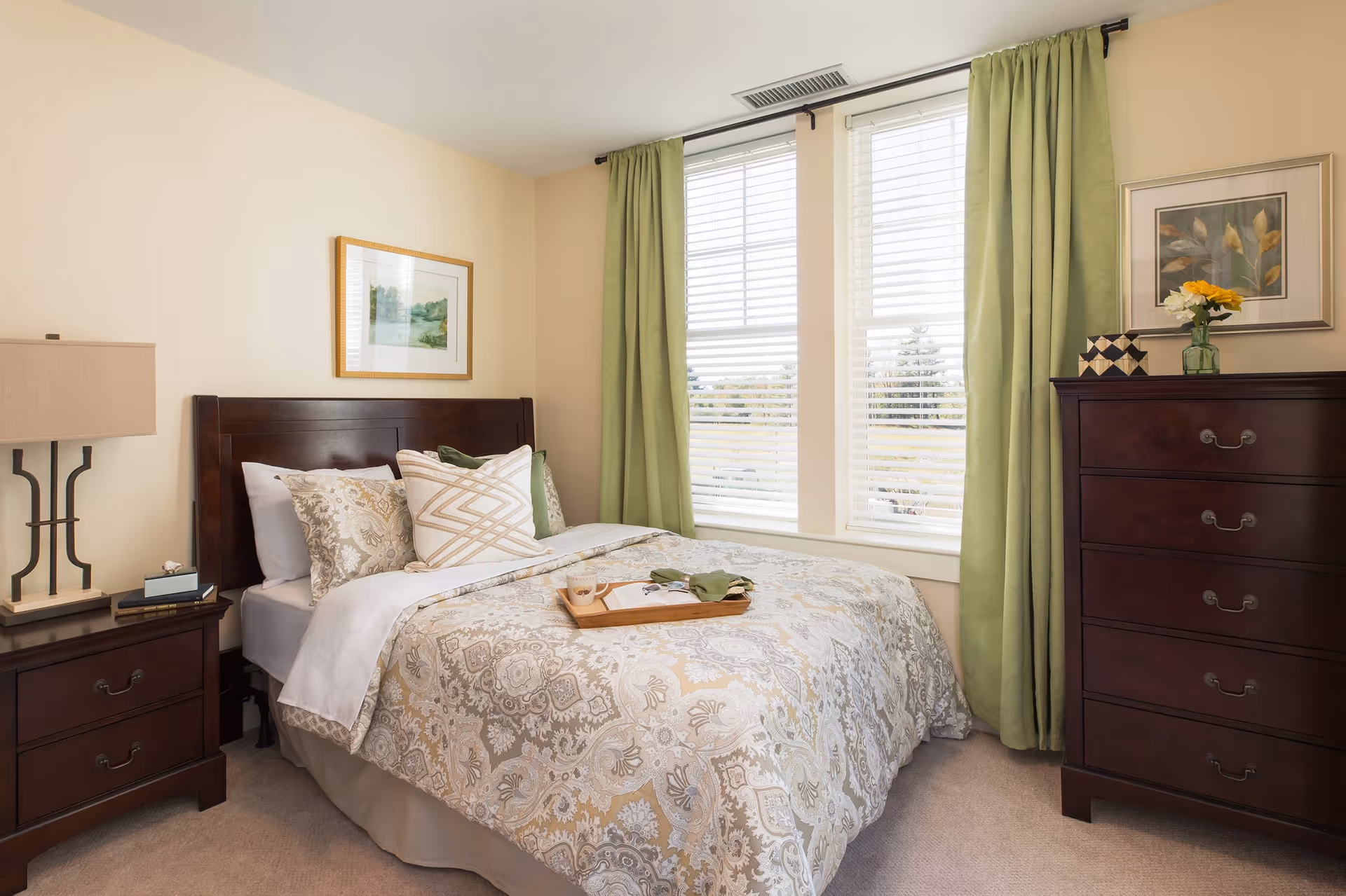 A neatly made bed with patterned bedding and multiple pillows in a bedroom. There is a wooden nightstand with a lamp and books on the left side of the bed, and a wooden dresser with a vase of flowers and decorative items on the right. Two large windows with white blinds and green curtains allow natural light into the room. A framed picture hangs on the wall above the bed and another above the dresser.