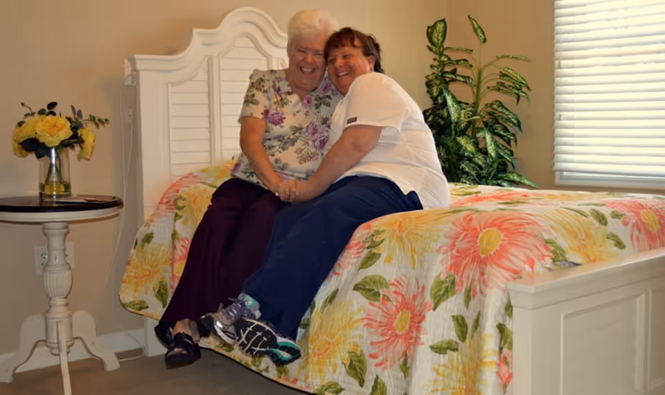 Two women sitting closely together on a bed with a floral bedspread in a cozy bedroom. One woman is elderly with white hair wearing a floral top and dark pants, and the other woman is wearing a white shirt and dark pants. There is a small round table with a vase of yellow flowers on the left side and a green leafy plant near the window with closed blinds on the right side.