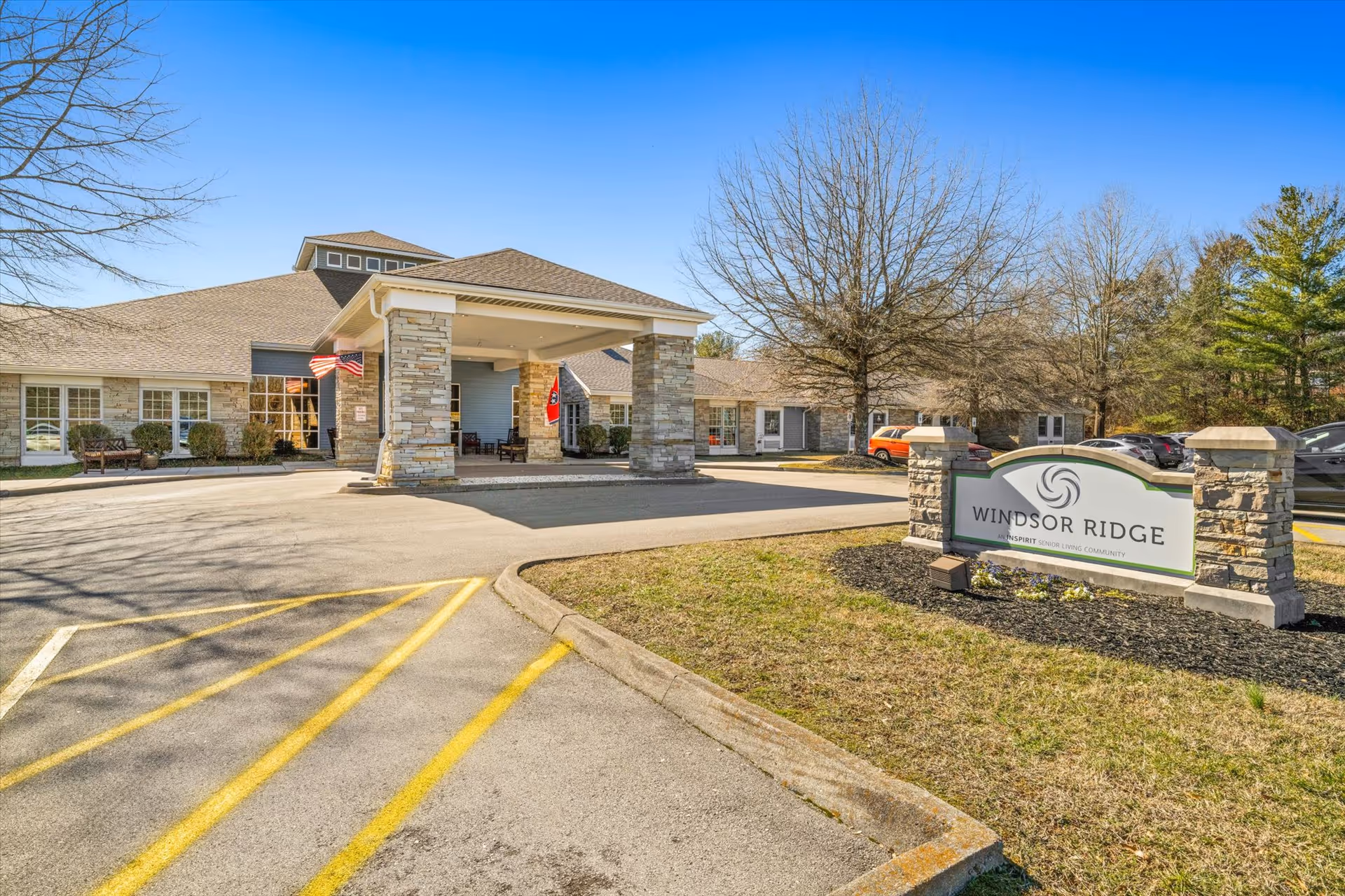 Exterior view of Windsor Ridge senior living community building with a covered entrance supported by stone pillars, an American flag and another flag hanging, a parking area with yellow lines, leafless trees, and a sign in the foreground that reads 'Windsor Ridge'.