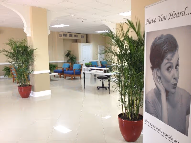 A bright and spacious interior area with shiny tiled floors, beige walls, and white ceiling tiles. There are several green potted plants in red pots placed around the space. Blue cushioned chairs with wooden frames are arranged in seating areas. A white table with a black office chair is also visible. On the right side, there is a large poster featuring a black and white photo of a woman with the text 'Have You Heard...' and additional smaller text below.