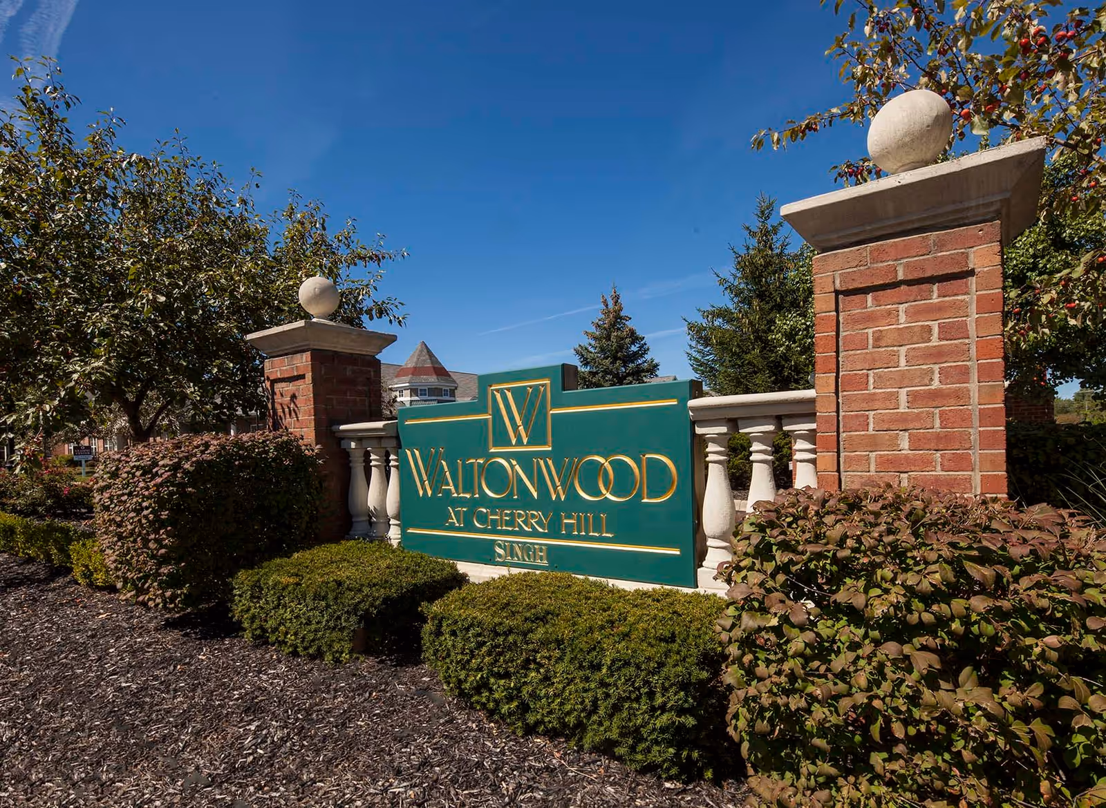 Outdoor view of the entrance sign for Waltonwood at Cherry Hill, surrounded by brick pillars, white balustrades, green bushes, and trees under a clear blue sky.