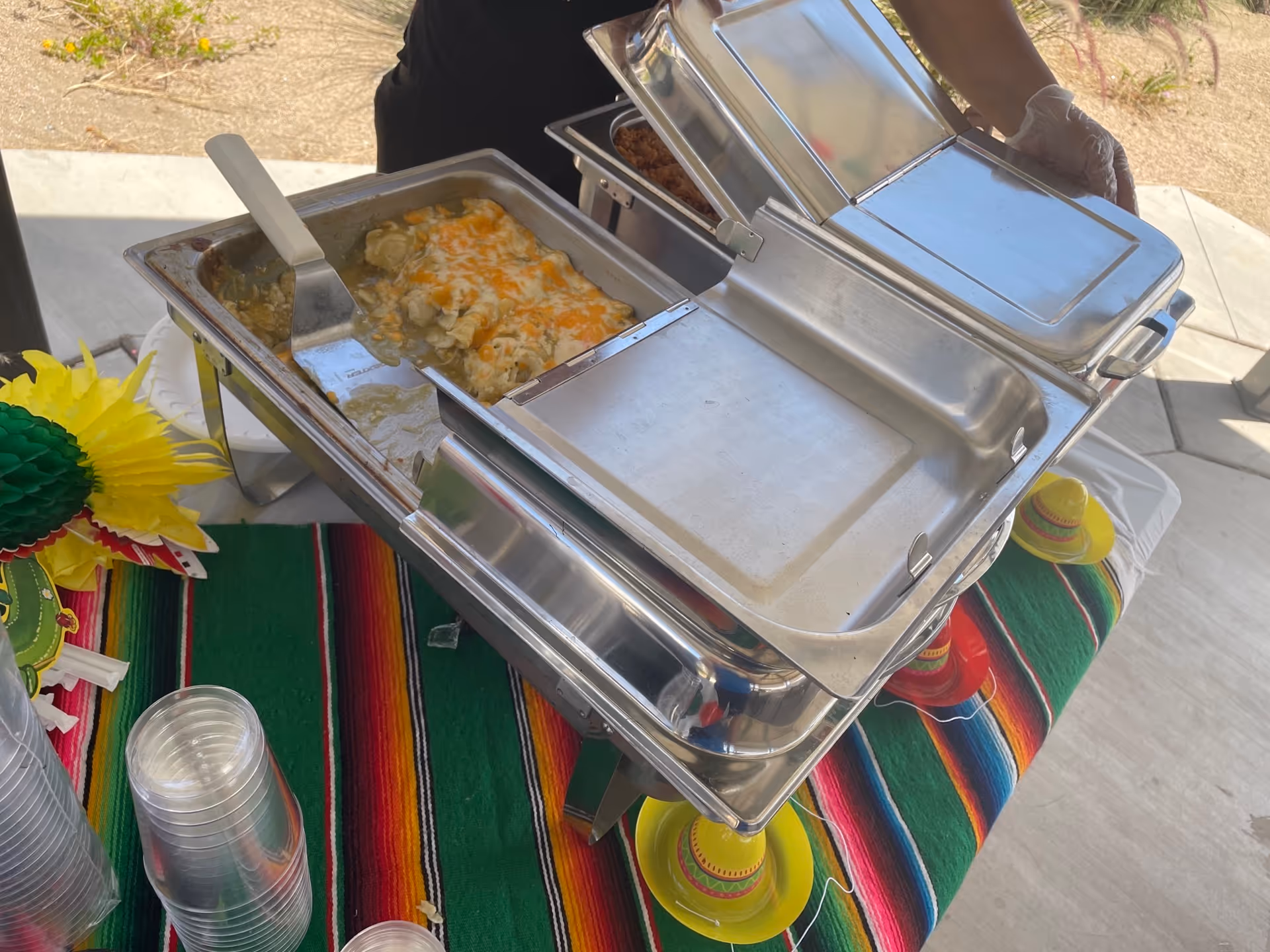 Chafing dishes with food and a serving spatula on a colorful striped tablecloth beside stacked plastic cups at an outdoor buffet.