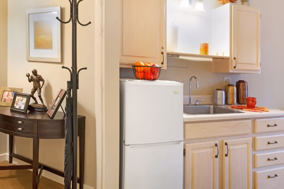 A small kitchen area with light wood cabinets, a white mini refrigerator topped with a fruit basket, a stainless steel sink, and various kitchen containers and a red mug on the counter. To the left, there is a dark wooden console table with framed photos and a bronze figurine, next to a black coat rack holding an umbrella.