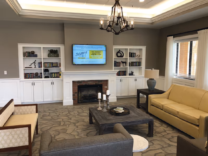 A cozy living room in a senior living facility featuring a yellow sofa, gray armchairs, a wooden coffee table with candles, built-in white bookshelves flanking a brick fireplace, and a wall-mounted TV above the fireplace. The room has a large window with white curtains and a chandelier hanging from the ceiling.