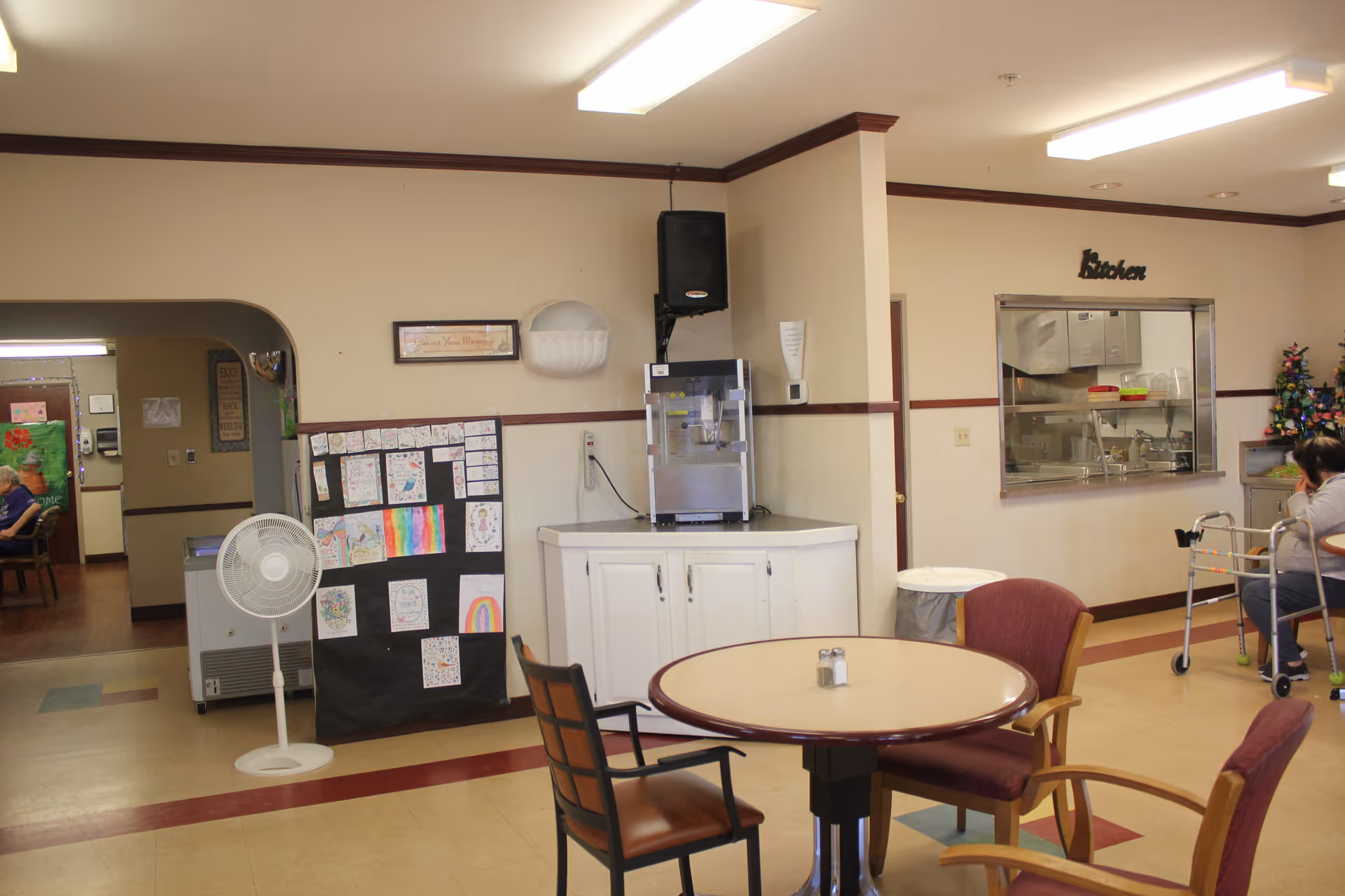 Interior view of a senior living facility common area with a round table and chairs, a water dispenser on a white cabinet, a bulletin board with children's drawings, and a serving window labeled 'Kitchen'. A person with a walker is seated near the serving window.