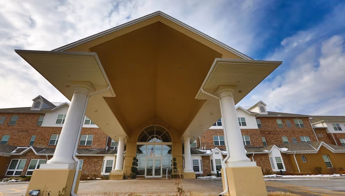 Front exterior view of a large brick and beige retirement community building with a covered entrance supported by white columns under a partly cloudy sky.