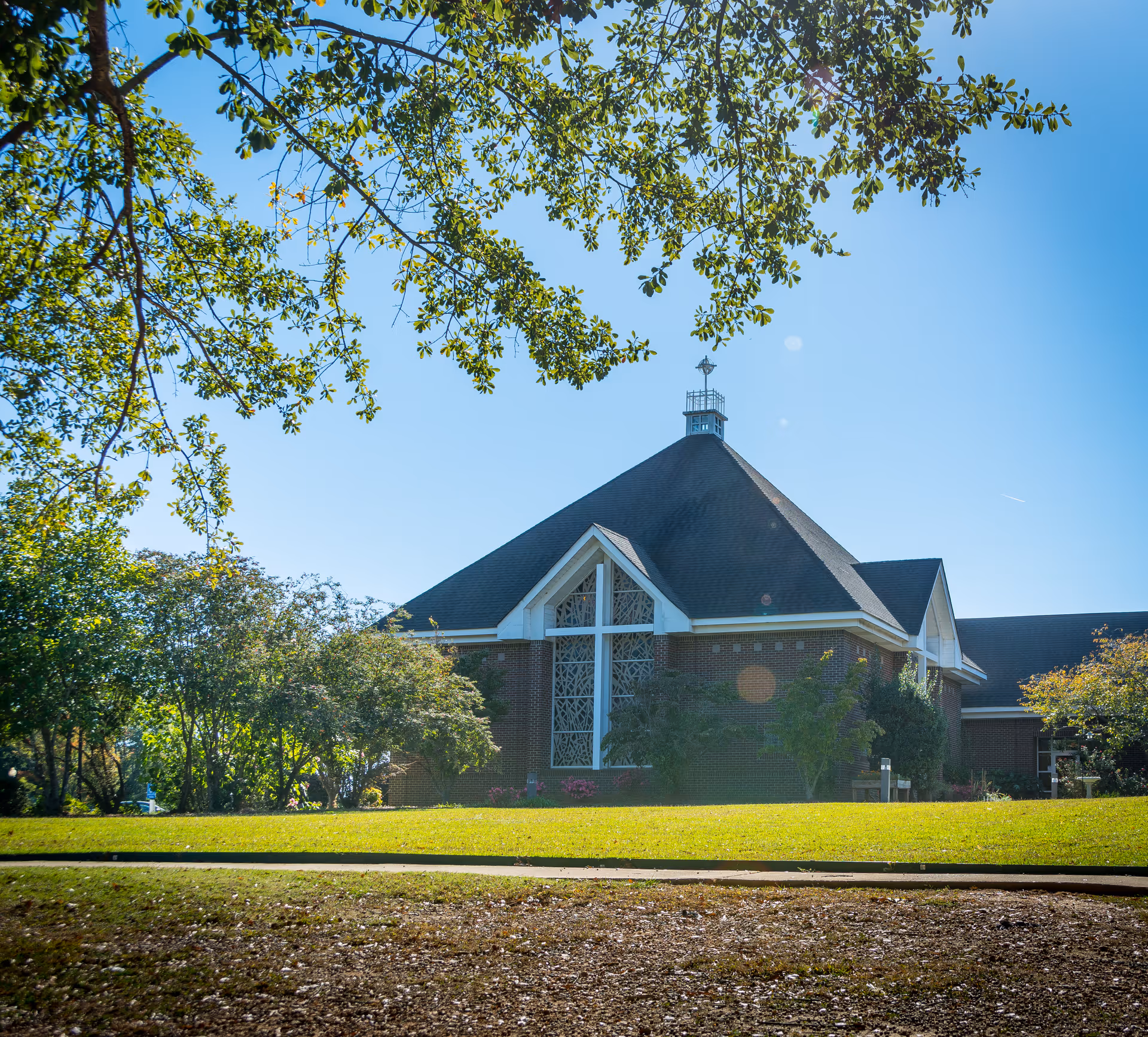 A brick building with a steep, dark roof and a large cross-shaped window, surrounded by green trees and grass under a clear blue sky.