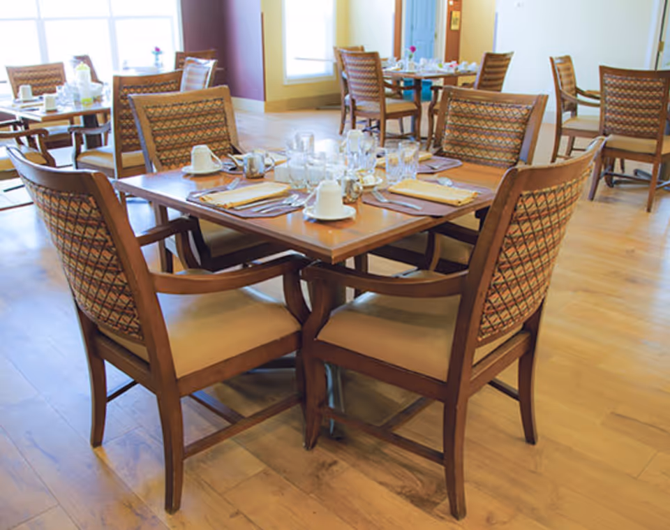 Dining area with a square wooden table set for four and patterned wooden chairs on a light wood floor.