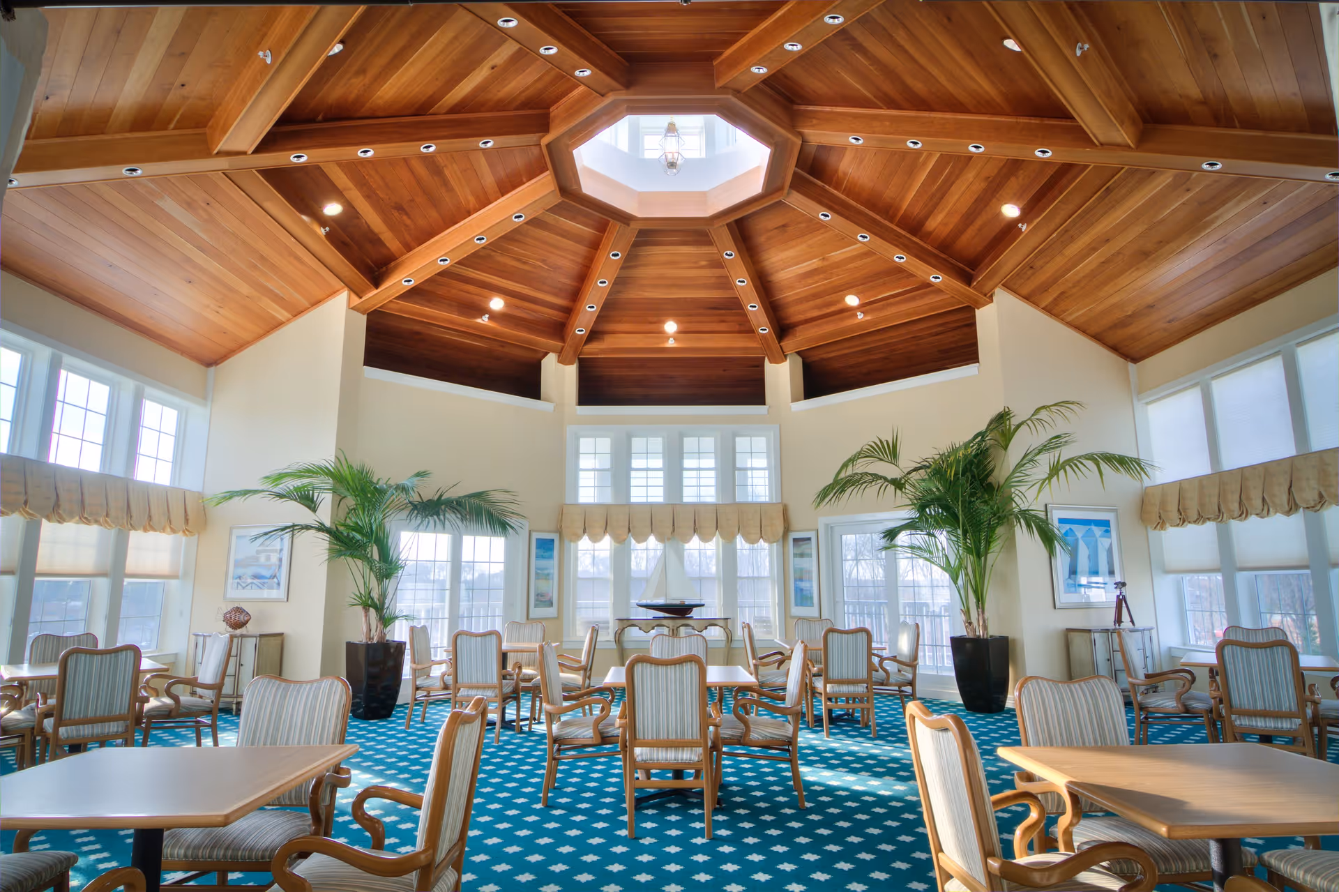 A spacious dining room with multiple wooden tables and cushioned chairs arranged neatly on a blue carpet with white patterns. The room features large windows allowing natural light to fill the space, two large potted plants, framed artwork on the walls, and a wooden octagonal ceiling with recessed lighting and a central skylight.