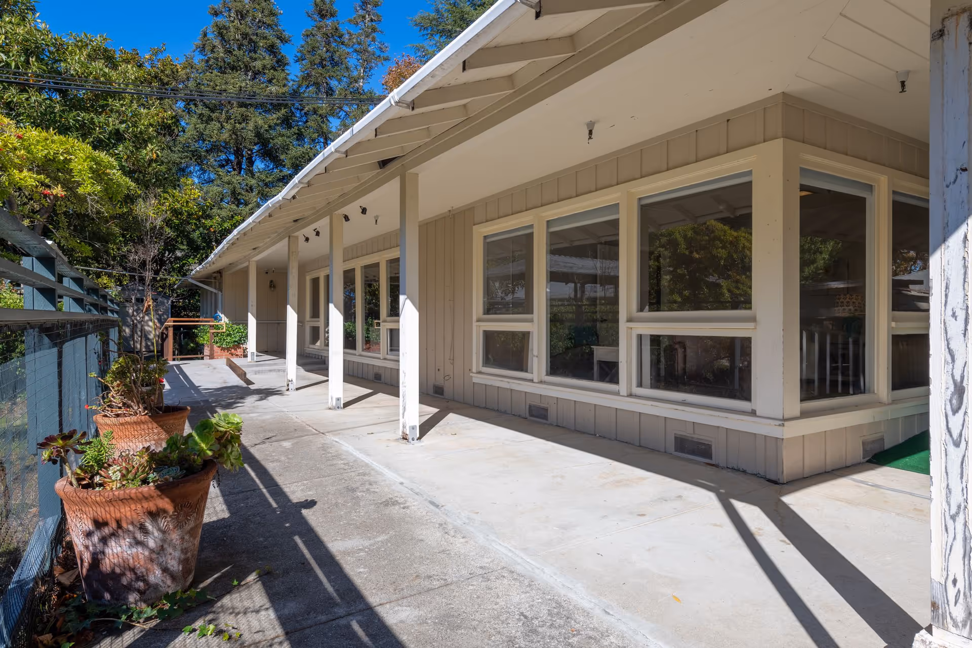 Outdoor covered walkway along the side of a building with large windows and several potted plants. Trees and a clear blue sky are visible in the background.