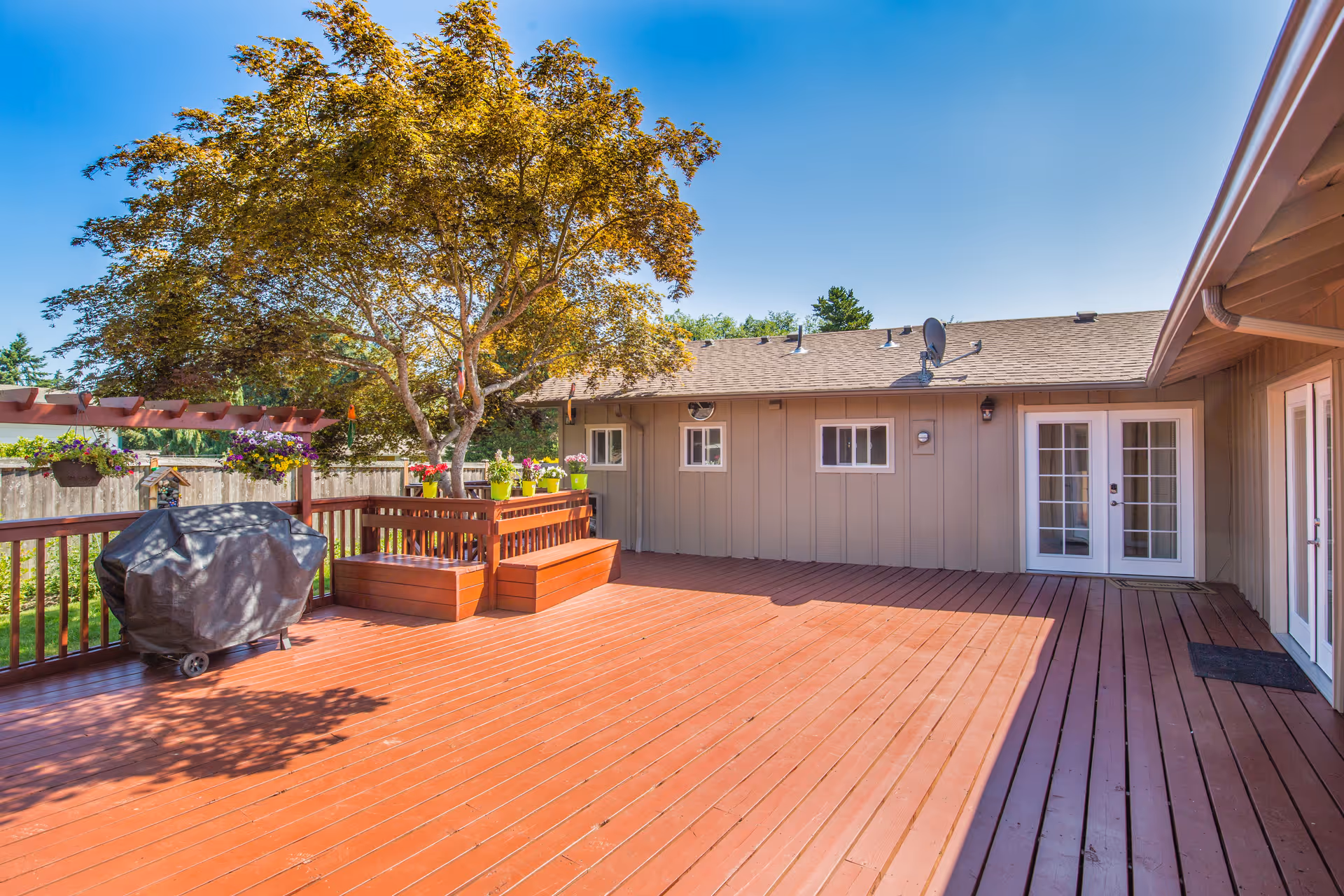 Spacious outdoor wooden deck area with a covered grill, built-in wooden benches, potted plants, and a tree providing partial shade. The deck is attached to a beige building with multiple windows and two sets of French doors under a clear blue sky.