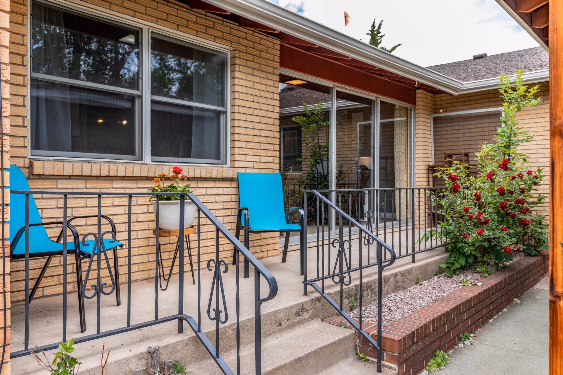 Small exterior patio with turquoise chairs, potted plants, metal railing, and sliding glass doors set against a brick building.