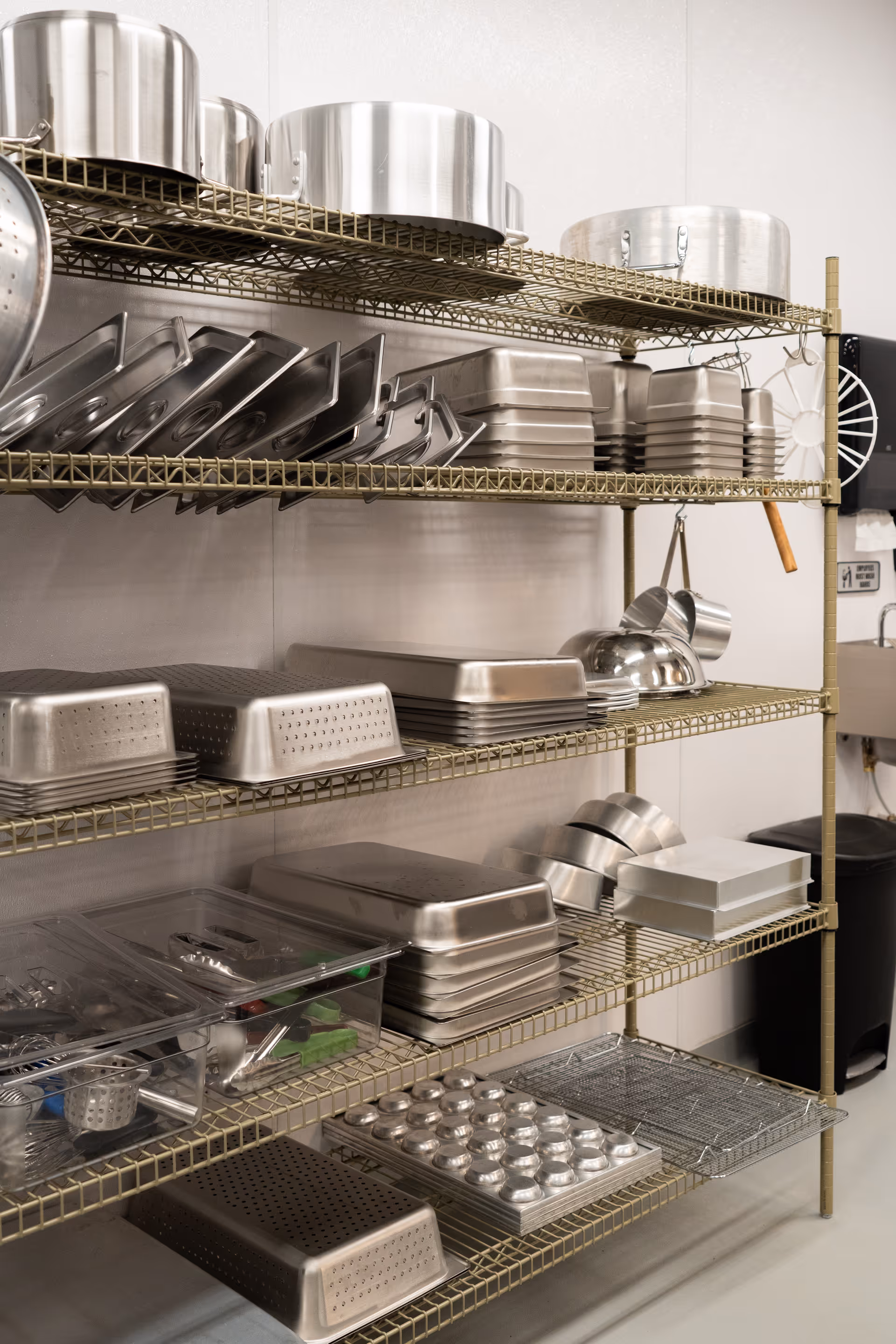 Wire shelving in a commercial kitchen stocked with stainless-steel pots, pans, baking trays, and utensils.