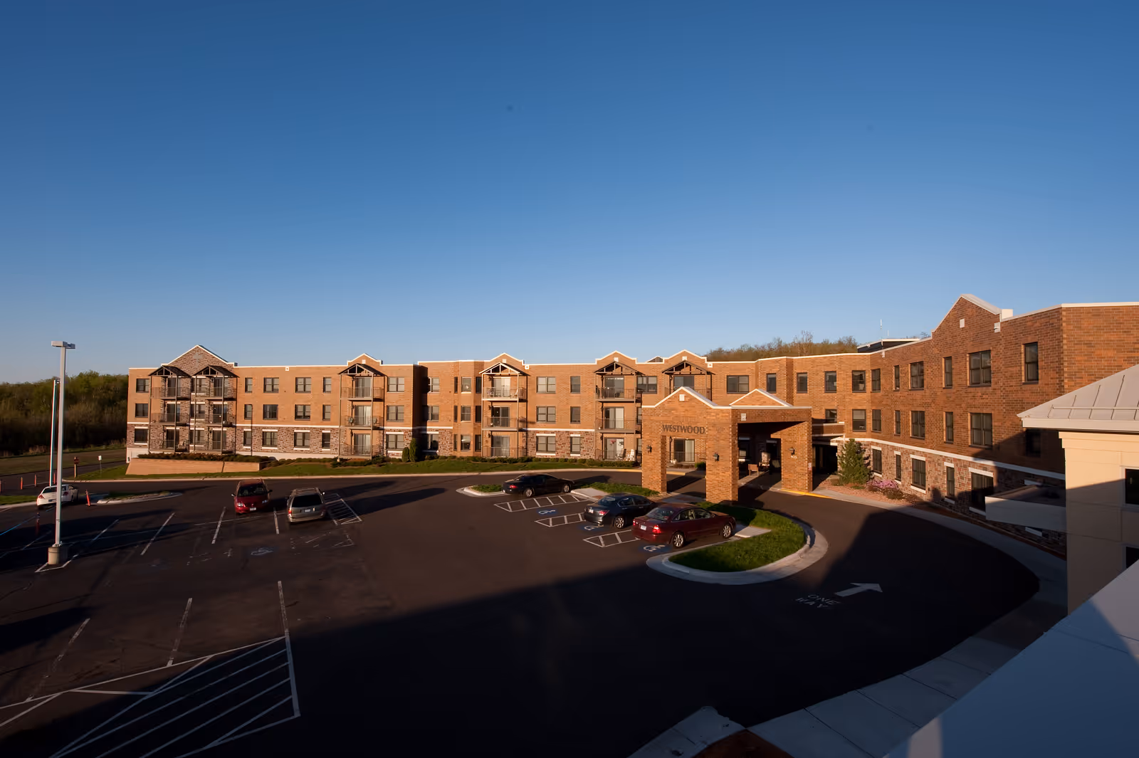 Brick three-story senior living building with balconies, a circular driveway and parking lot under a clear blue sky.