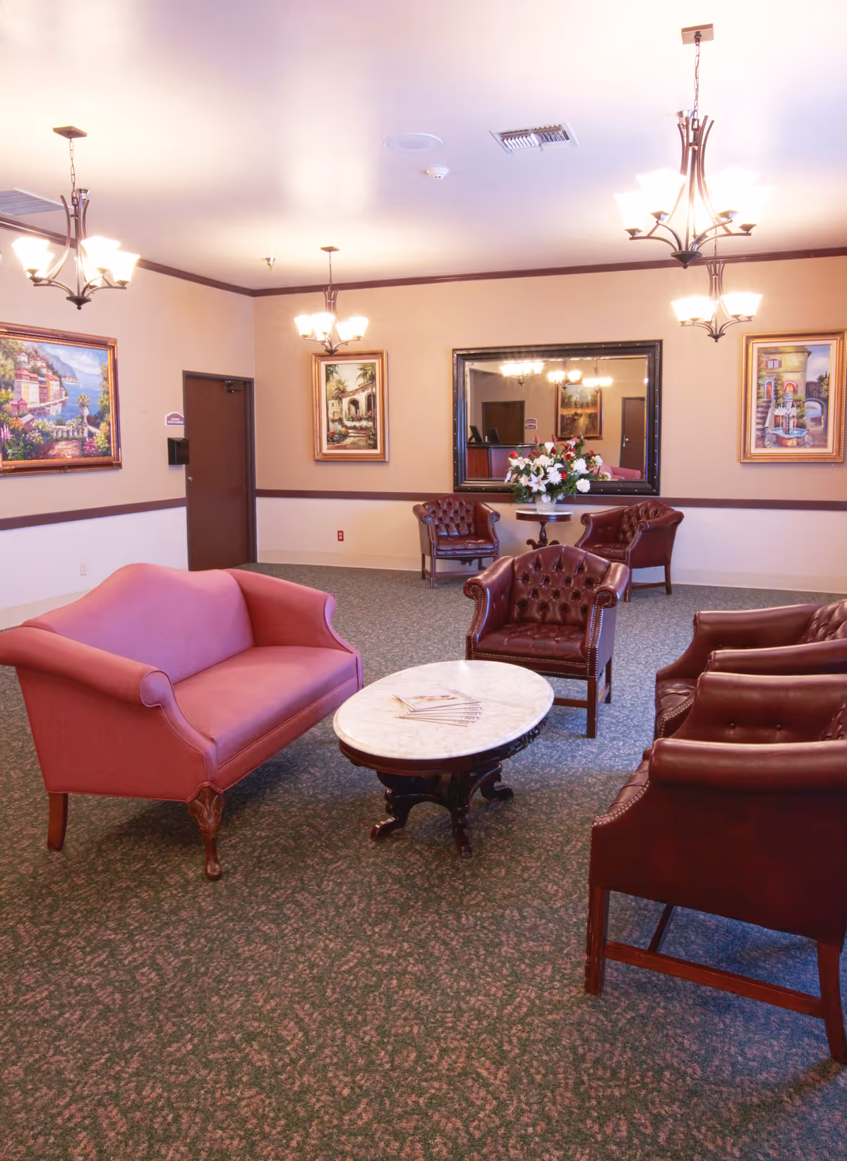 A cozy sitting area in a senior living facility with a pink upholstered loveseat, four brown leather armchairs, and a round marble coffee table in the center. The room has patterned carpet, beige walls with dark wood trim, three framed paintings, a large wall mirror, and three ceiling light fixtures with multiple bulbs.