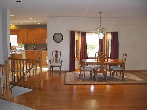 Open dining area with wooden table, chairs, chandelier, and kitchen in background.