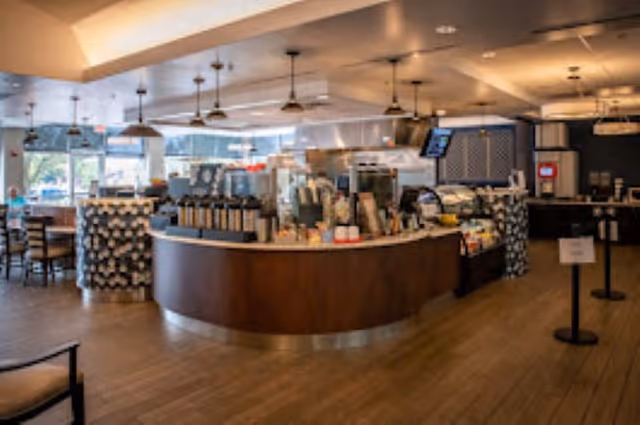 Interior view of a modern dining area with a curved counter serving coffee and food items. The space has wooden flooring, pendant lights hanging from the ceiling, and tables and chairs arranged near large windows letting in natural light.