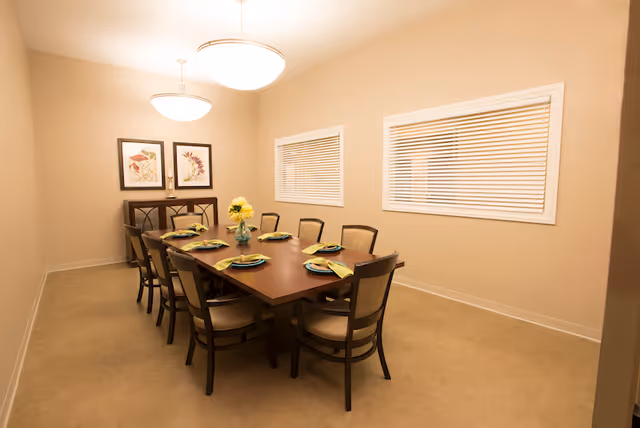 A dining room with a rectangular wooden table set for eight, chairs, pendant lights, a floral centerpiece, and beige walls with two windows.