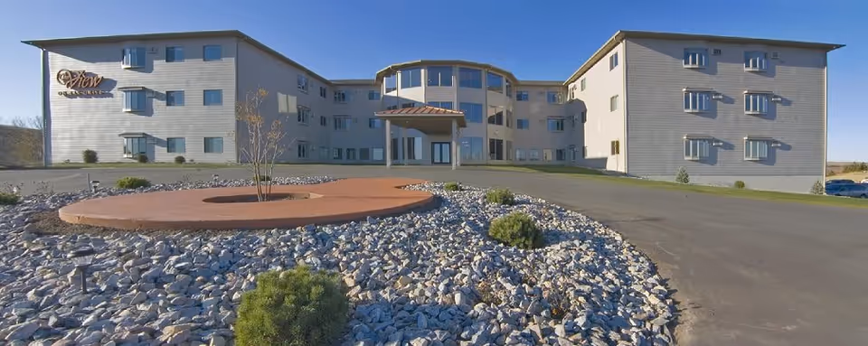 Front exterior view of a three-story senior living facility building named The View on Elk Drive, with a circular driveway and landscaped rock garden with small bushes and a young tree in front.