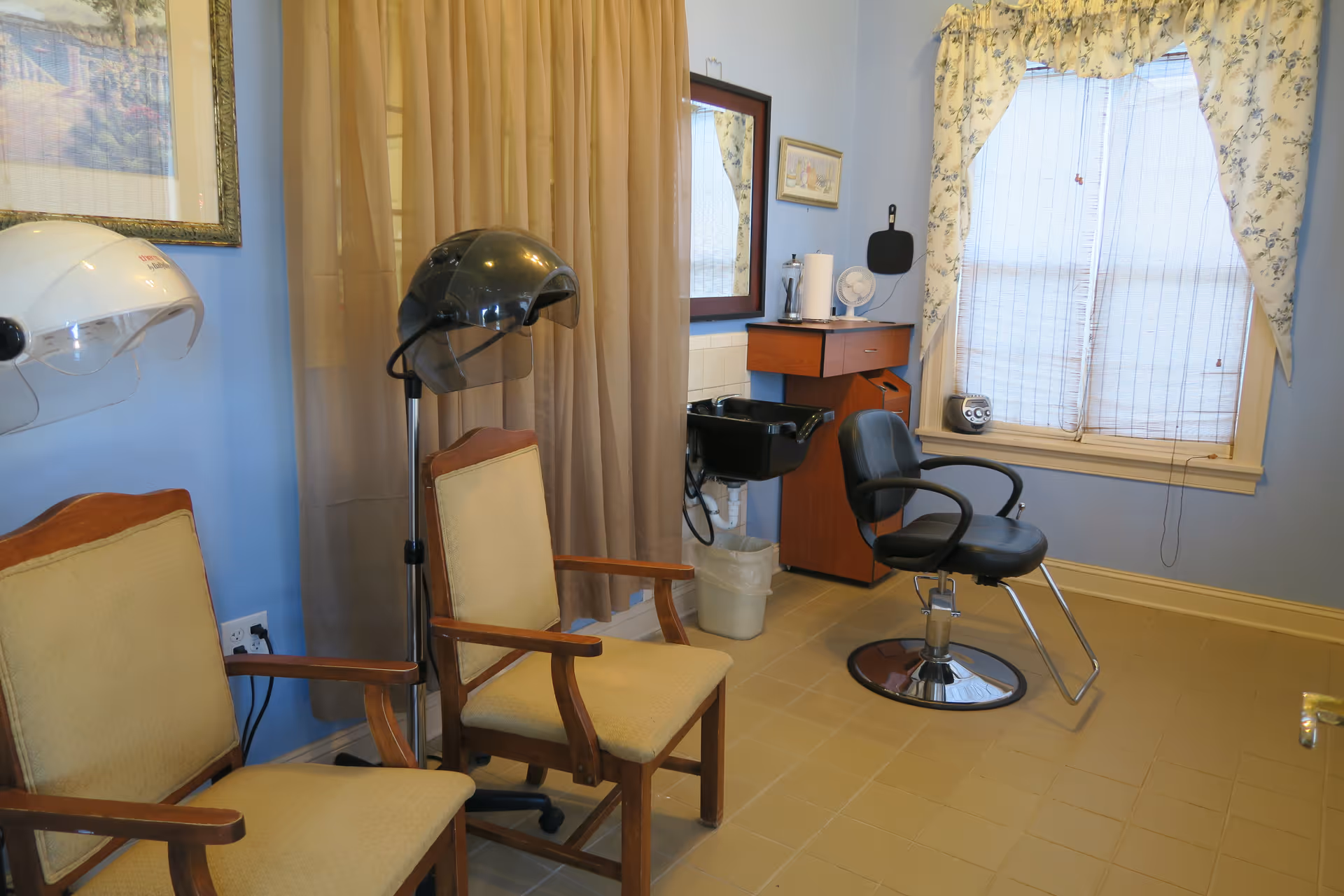 Interior of a small hair salon area with two beige cushioned wooden chairs, a black salon chair in front of a black hair washing sink, two hair dryers mounted on stands, a window with floral curtains, a mirror, and a small wooden cabinet with a paper towel holder and a fan on top.