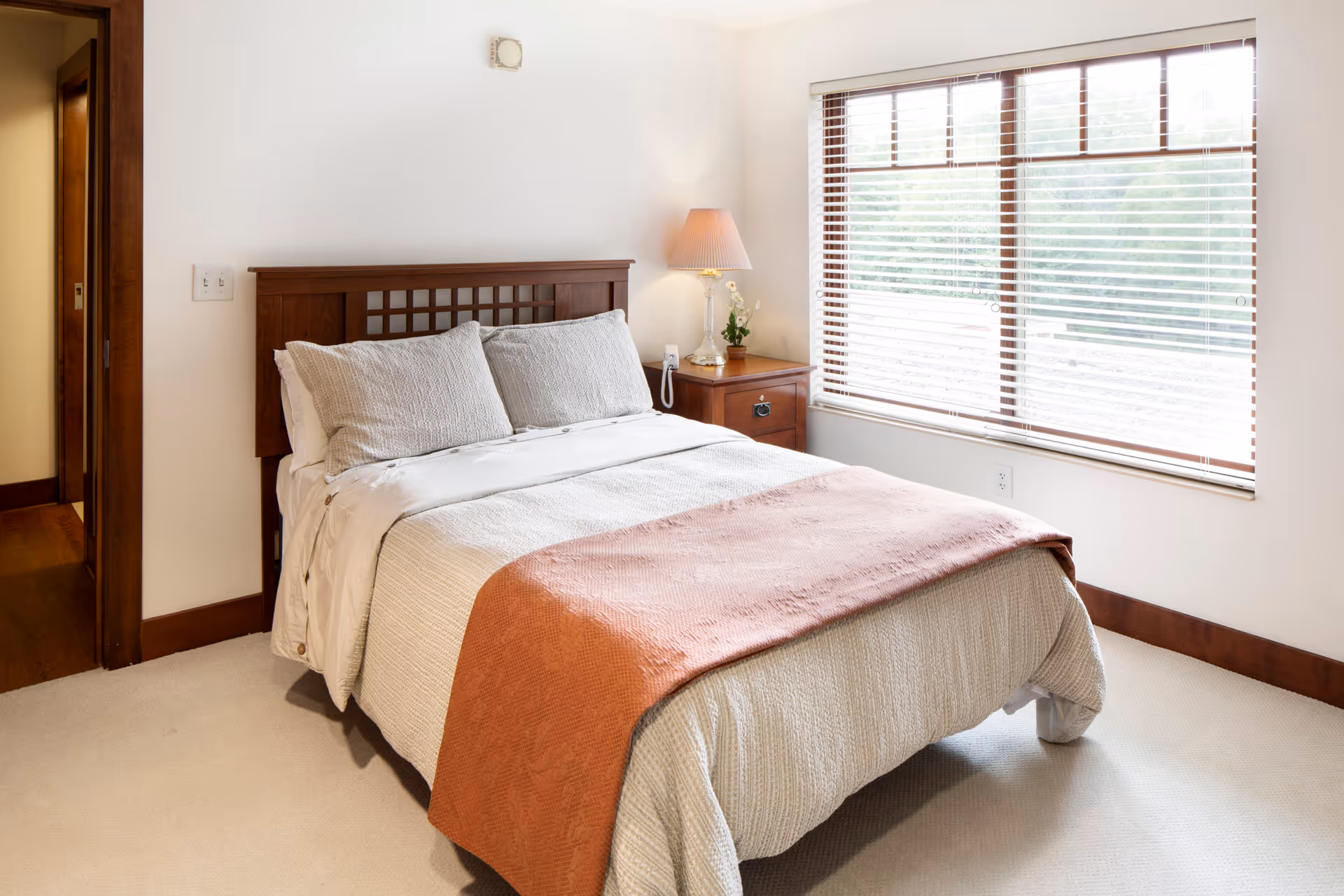Bright bedroom with a made bed, wooden headboard, nightstand with a lamp and vase, and a large window with blinds.