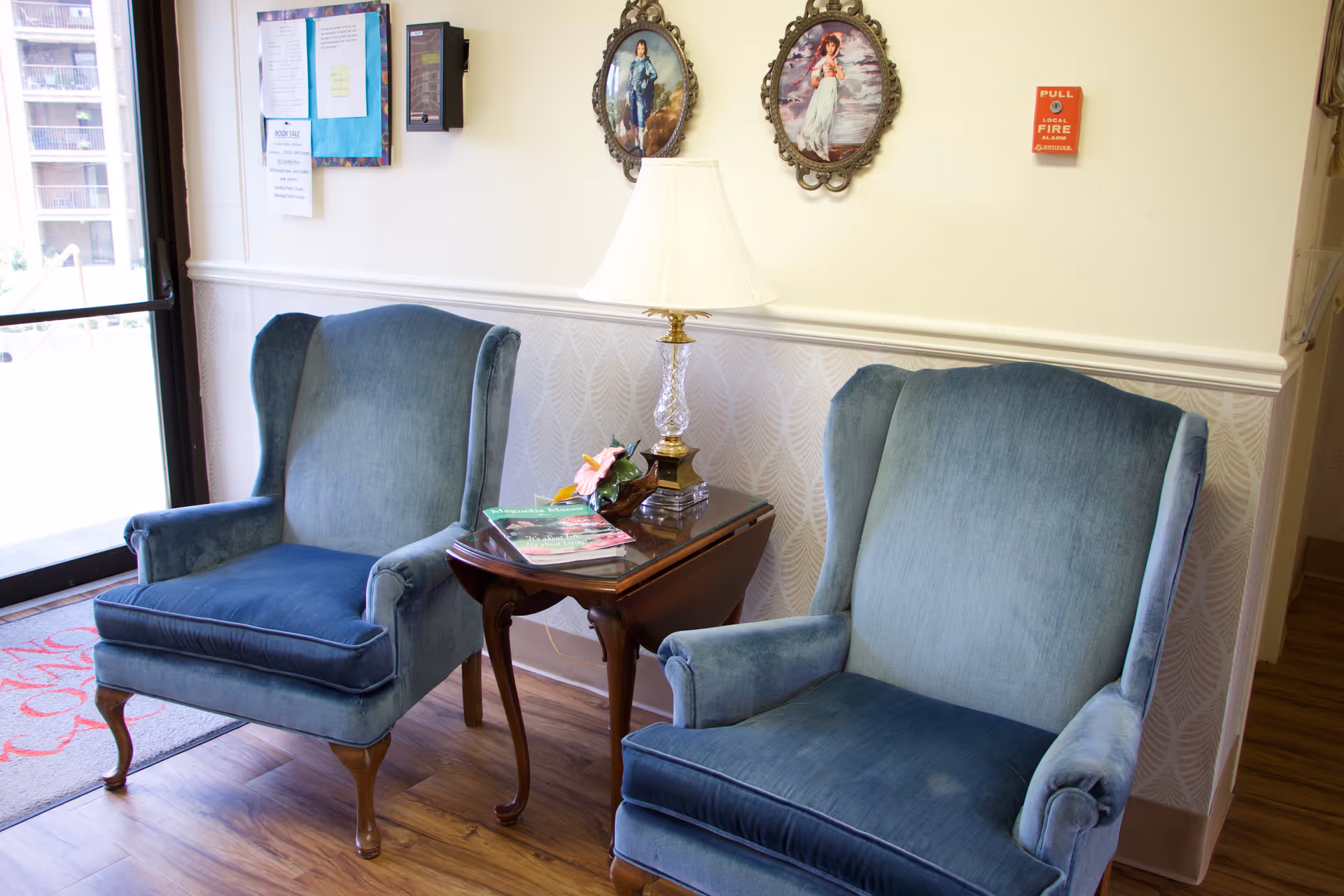 Two blue upholstered armchairs with wooden legs positioned on either side of a small wooden side table with a glass lamp and some magazines on it. The setting appears to be a cozy indoor seating area with framed pictures on the wall and a glass door to the left.