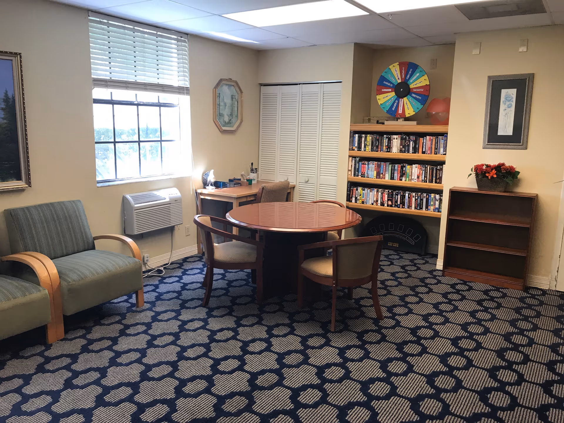 A cozy room with patterned carpet, two green armchairs, a round wooden table with three chairs, a small desk by a window with blinds, a bookshelf filled with books and a colorful prize wheel on top, and a small empty wooden shelf with a flower pot on it.