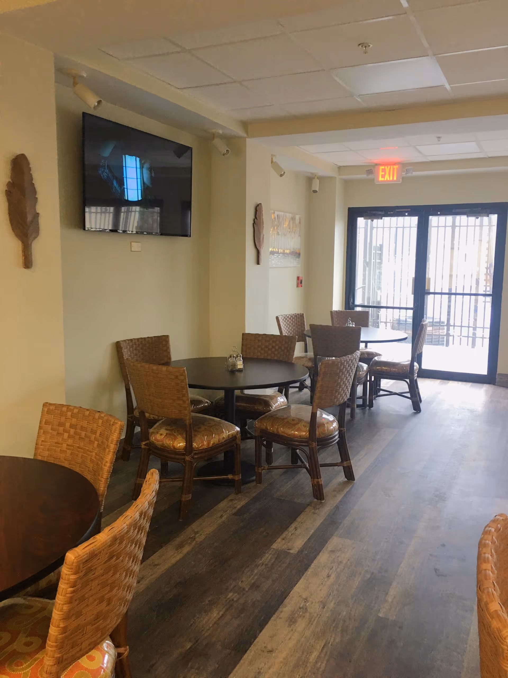 Interior view of a senior living facility dining area with round tables and wicker chairs arranged neatly. A flat-screen TV is mounted on the wall, and there is an exit door with glass panels letting in natural light.