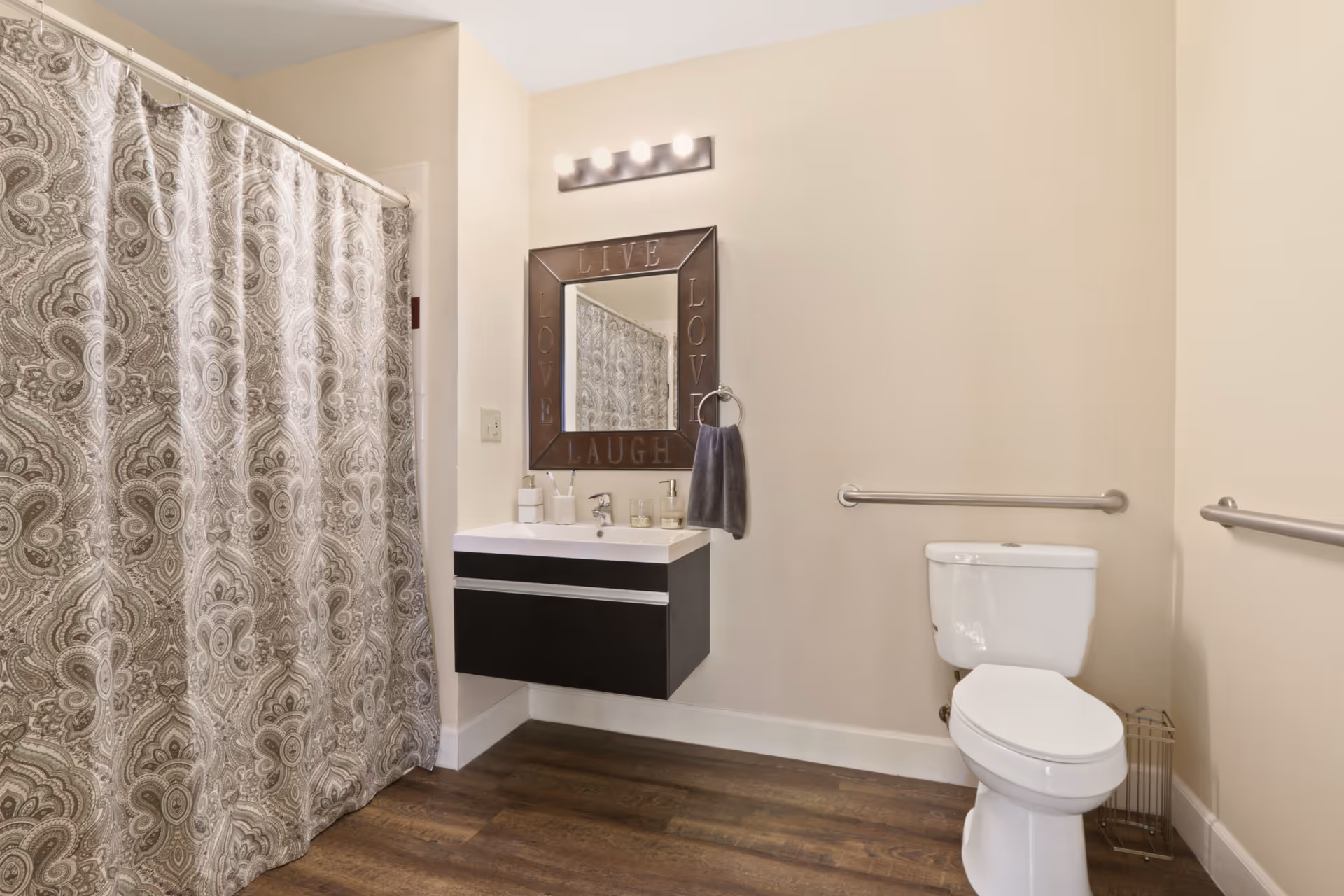 A bathroom with a patterned shower curtain on the left, a floating vanity with a sink and toiletries, a mirror with the words 'LIVE LOVE LAUGH' around its frame, a towel ring with a dark towel, a toilet with grab bars on the walls, and wood-look flooring.