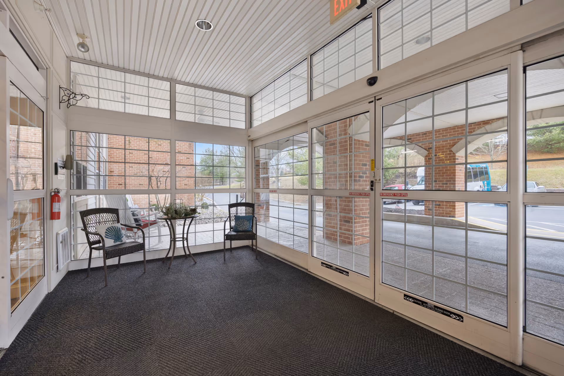 A bright enclosed entryway with large glass windows and automatic sliding glass doors. Two black chairs with decorative pillows and a small round glass table with a plant are placed near the windows. The exterior shows a brick building and a parking area with vehicles visible outside.