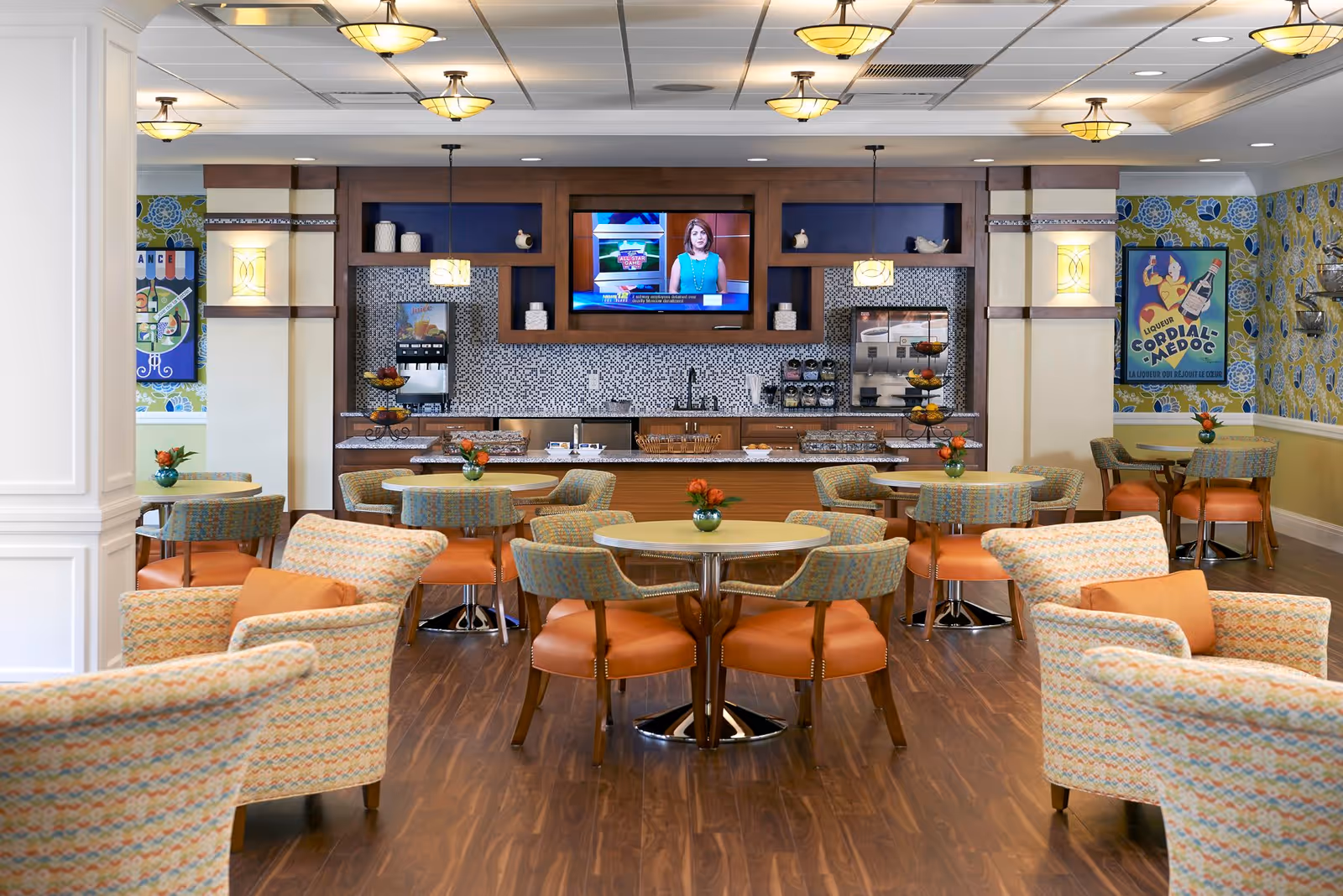 A brightly lit dining area with multiple round tables and cushioned chairs featuring orange seats and patterned backs. The room has wood flooring and a counter area in the back with a mosaic tile backsplash, beverage dispensers, and a mounted flat-screen TV displaying a news anchor. The walls have colorful floral wallpaper and framed vintage-style posters. Small flower arrangements are placed on each table.