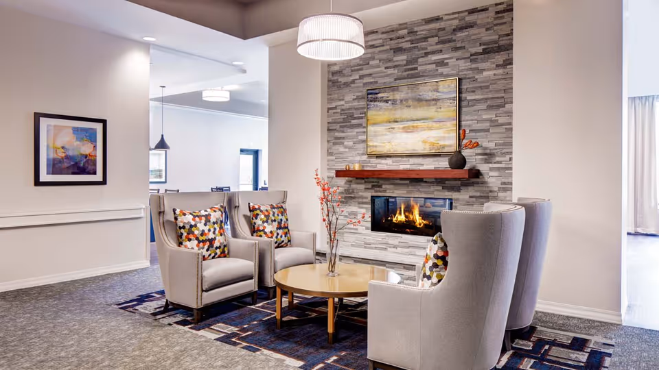 A cozy sitting area with four gray upholstered armchairs arranged around a round wooden coffee table. The chairs have colorful patterned pillows. Behind the chairs is a modern fireplace set in a gray stone accent wall with a wooden mantel holding a black vase with orange flowers and a small candle. Above the mantel is a framed abstract painting. The room has soft lighting from a ceiling fixture and an open doorway leading to another room.