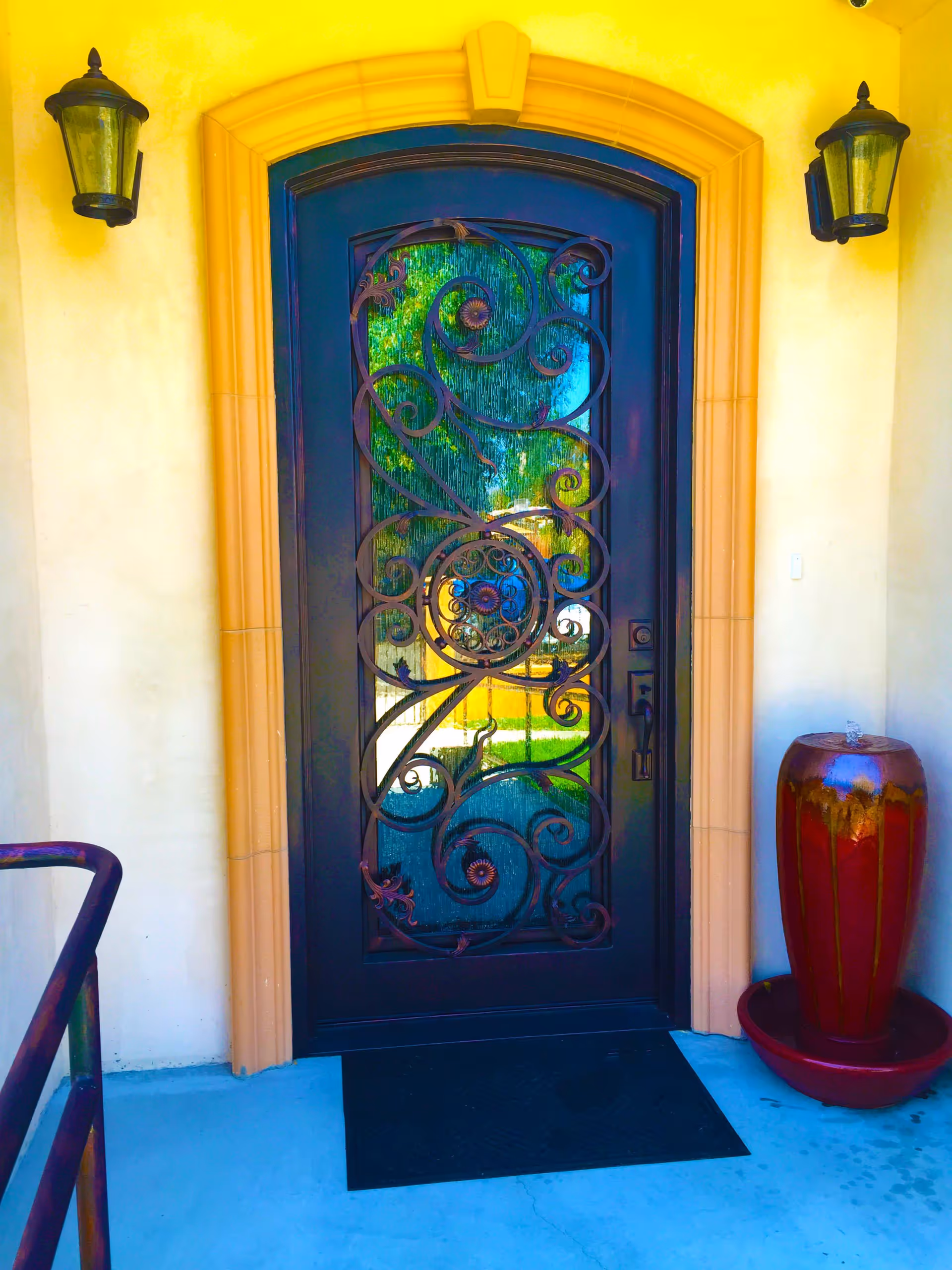 A decorative black metal front door with intricate scrollwork and textured glass panels, set in a yellow stucco wall with a tan archway trim. Two black wall-mounted lantern lights flank the door. To the right of the door is a tall red ceramic water fountain with water flowing from the top into a basin below. A black doormat is placed in front of the door on a light blue concrete floor.